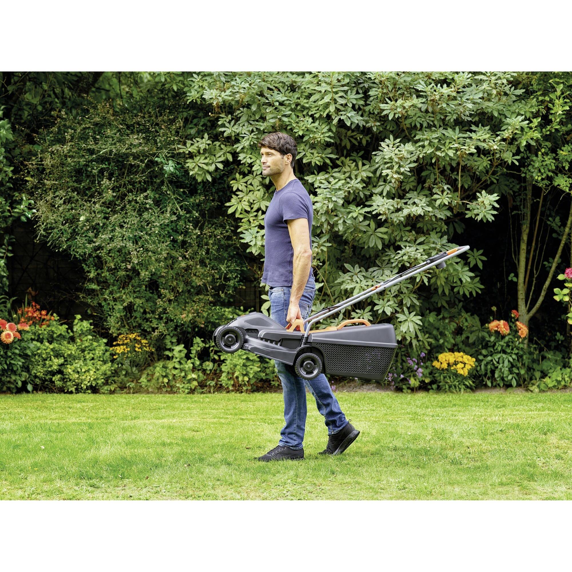 A person is carrying a lawnmower across a green lawn. Bushes and flowers are situated in the background.