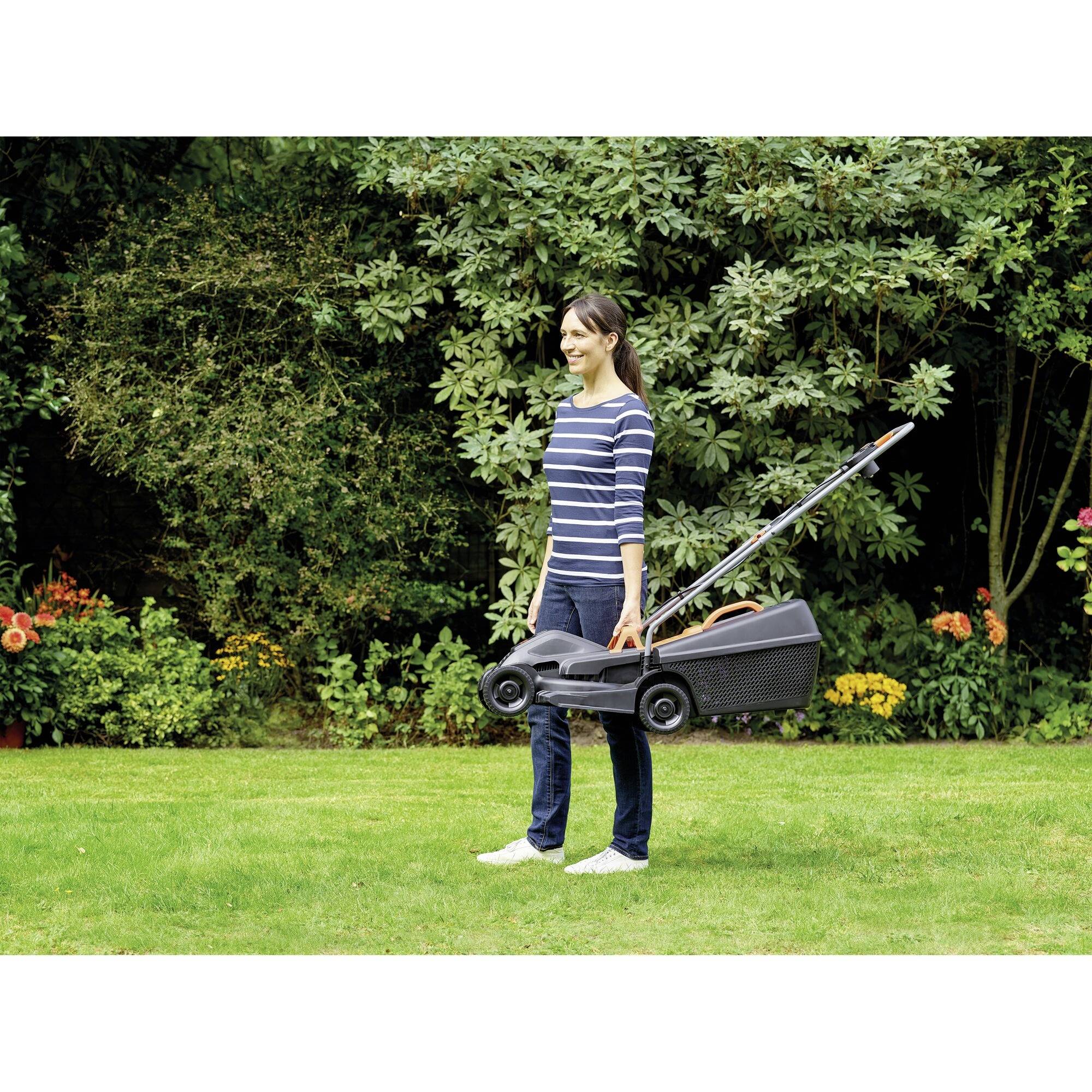 A person is standing with an upright lawnmower in a garden. Bushes and flowers can be seen in the background.