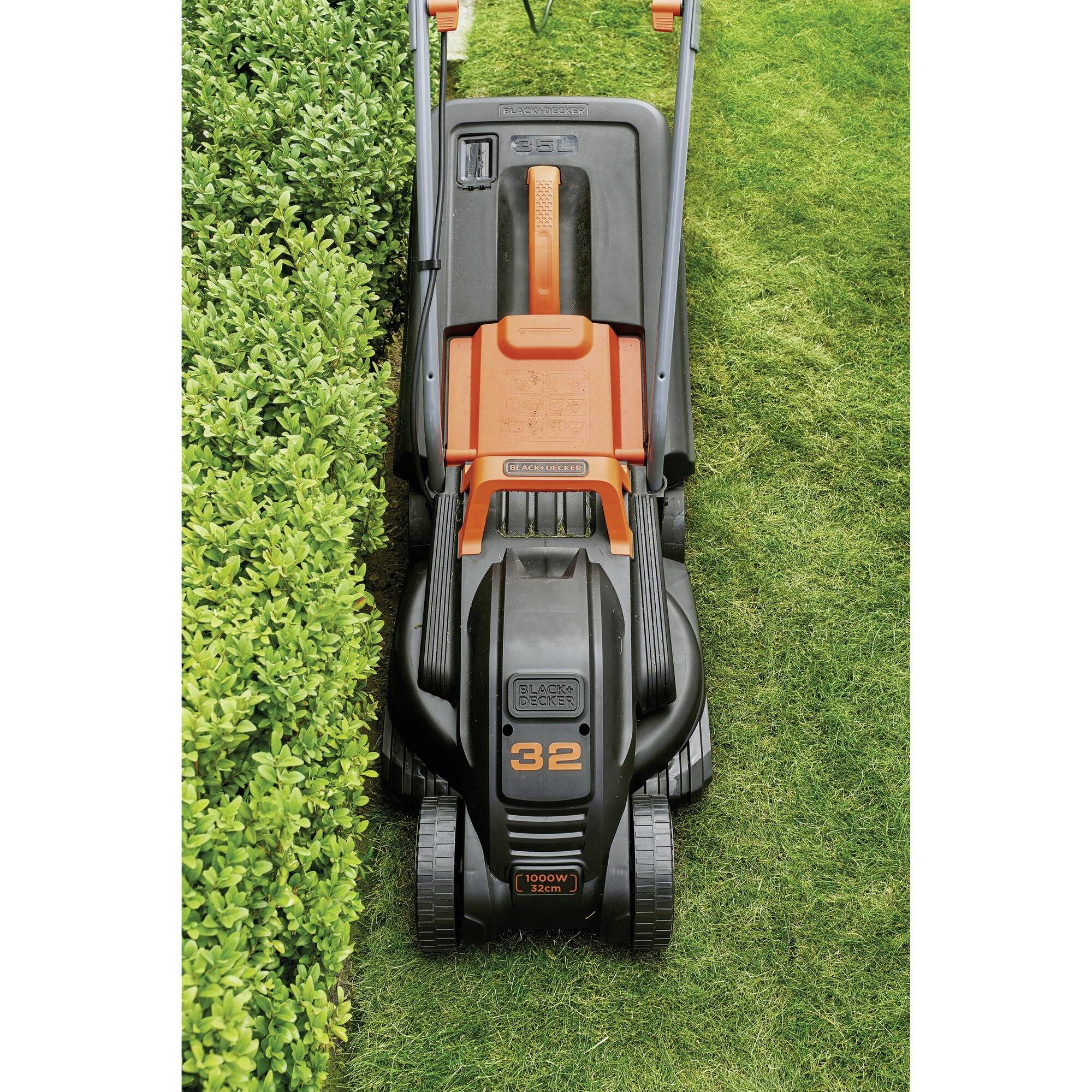 A lawn mower is cutting a strip of grass next to a hedge. The device is in black and orange, with the number '32' on the front.