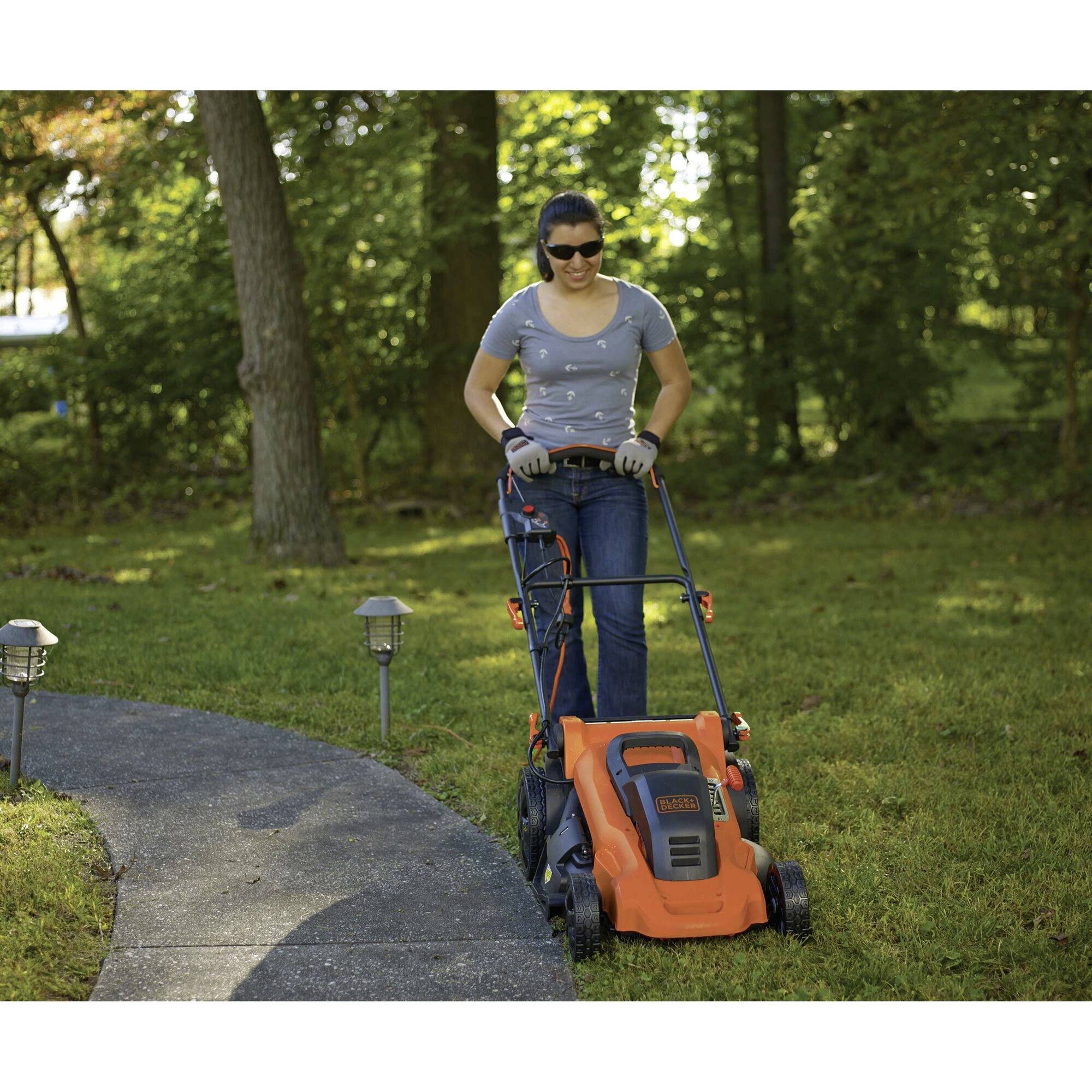 A person is mowing the lawn in the garden with an orange lawnmower, wearing sunglasses and gloves. Trees are visible in the background.