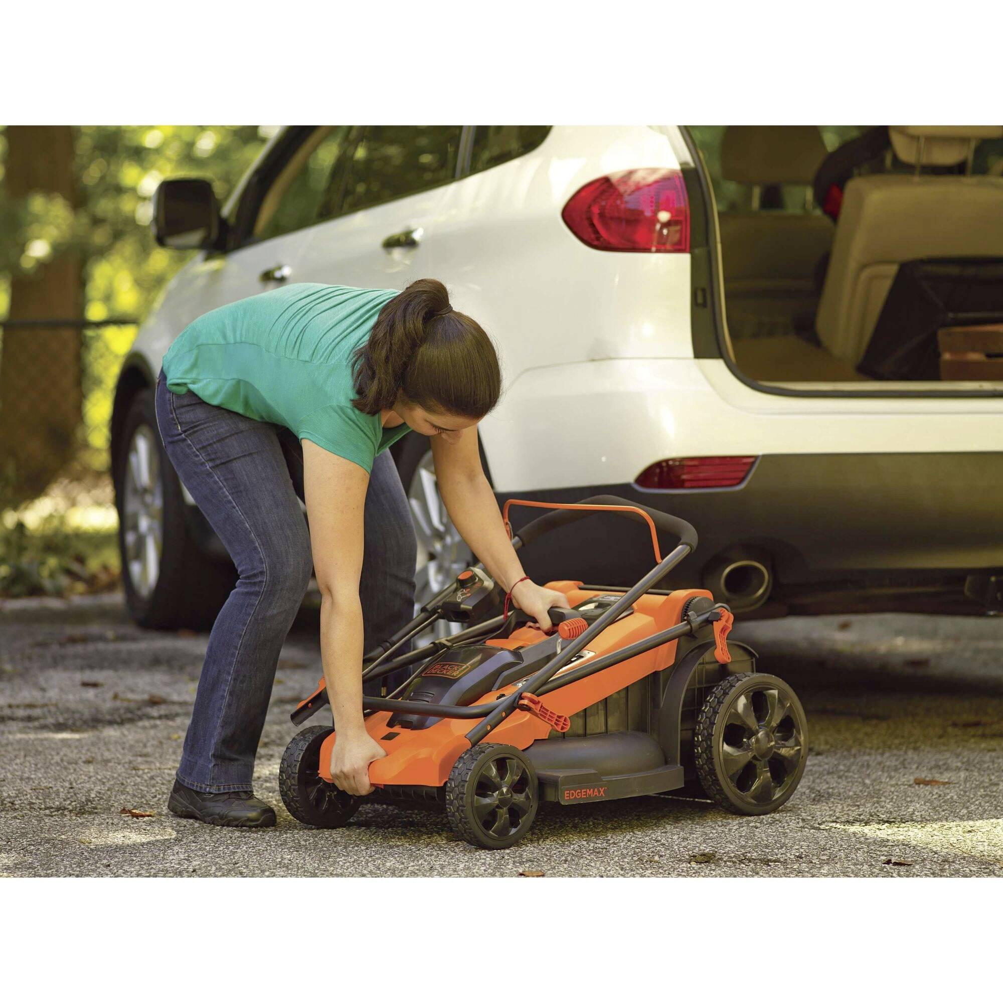 A person is unfolding a foldable lawnmower next to a car park. A white car is parked in the background.
