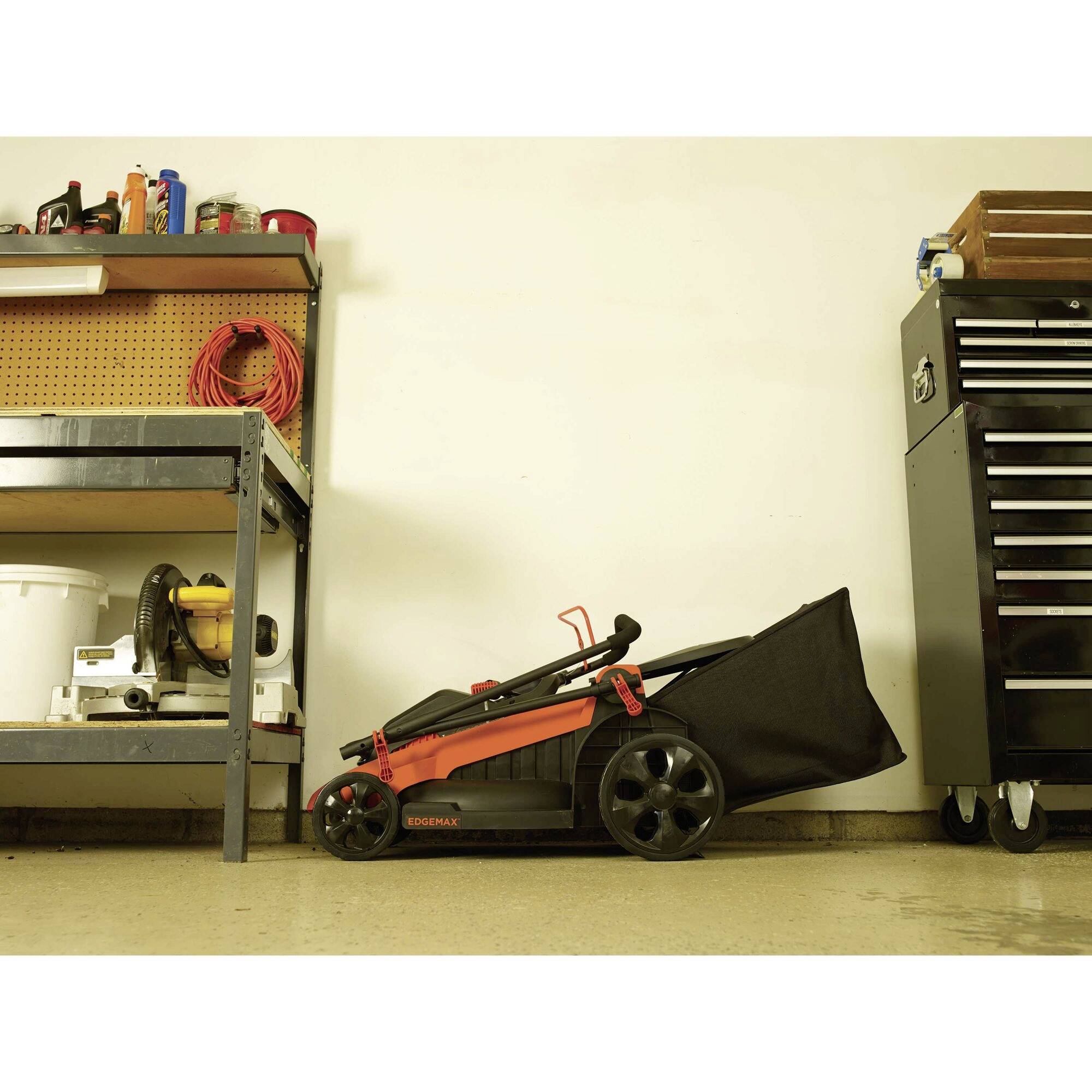A red and black lawnmower is standing on the floor of a workshop. To the right is a tool cabinet, and to the left is a table with tools and cables.