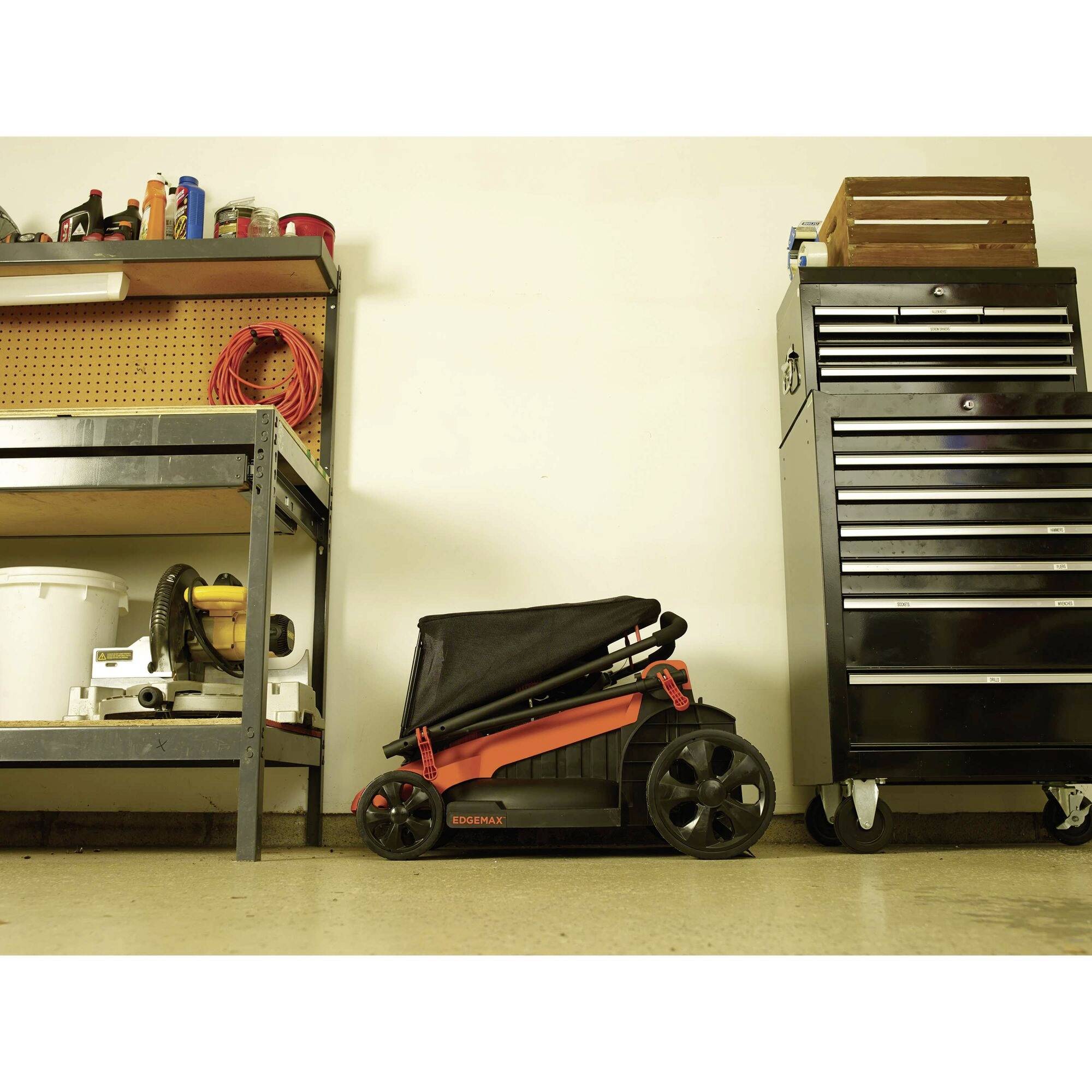 'Garage with tools and equipment: Workbench with electric saw, red cable reel overhead, black toolbox on wheels, orange pushchair in the middle.'