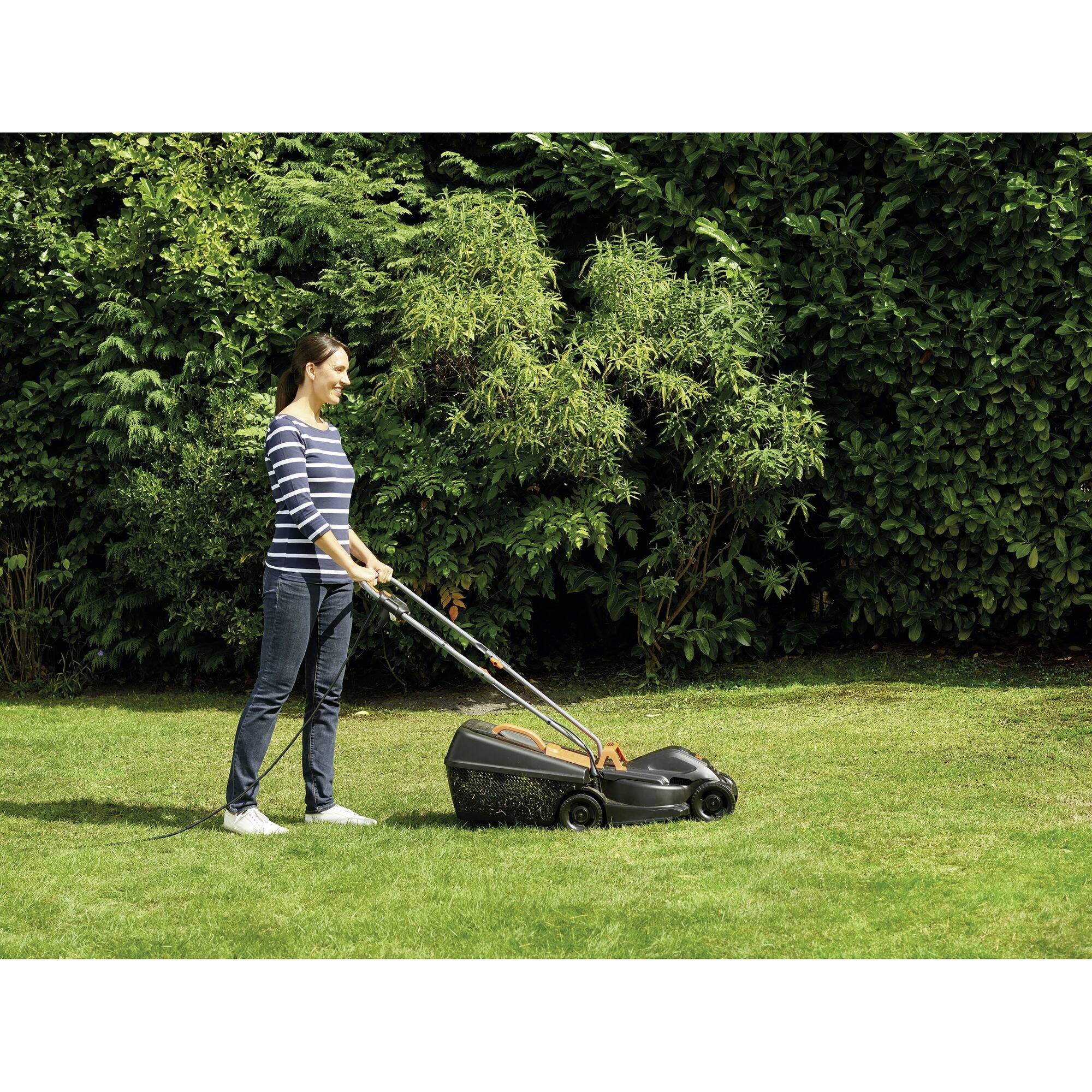 A person is mowing the lawn with an electric lawnmower in a green garden, surrounded by dense shrubbery in the background.