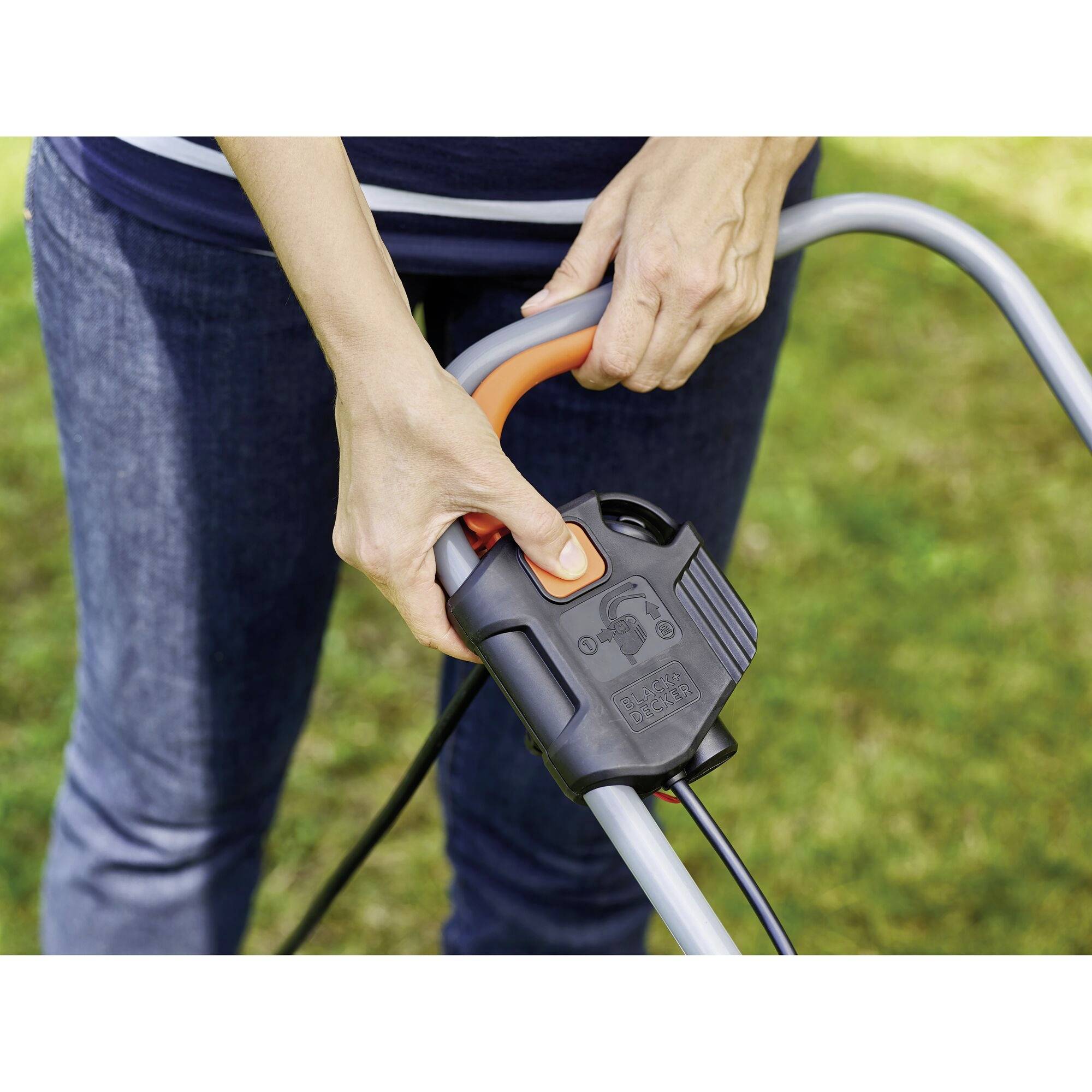 A person is operating an electric lawnmower. Hands are gripping the handle with control element, ready to start. Lawn visible in the background.