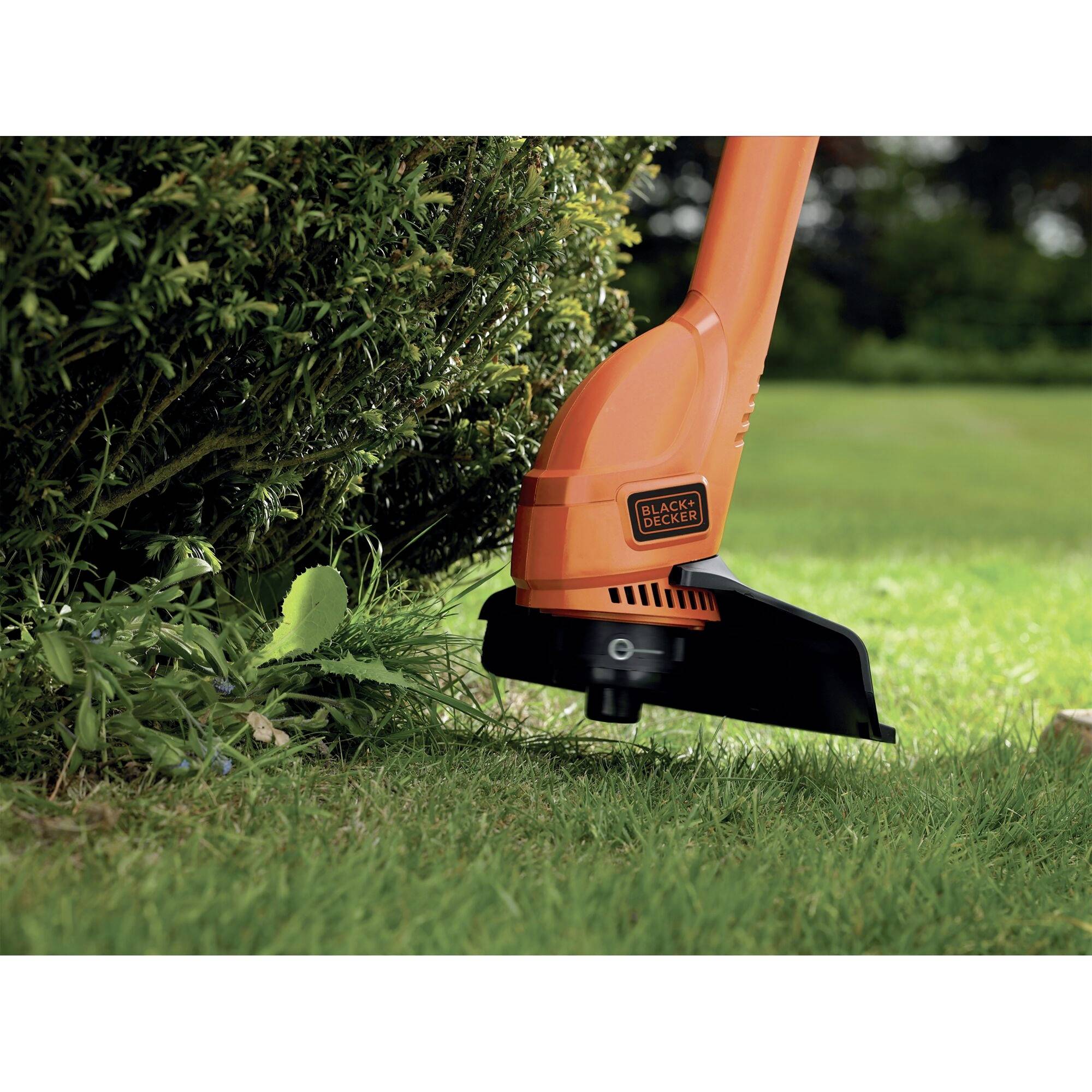 An orange strimmer is trimming grass edges beside a bush in a garden. In the background, a lawn area is visible.