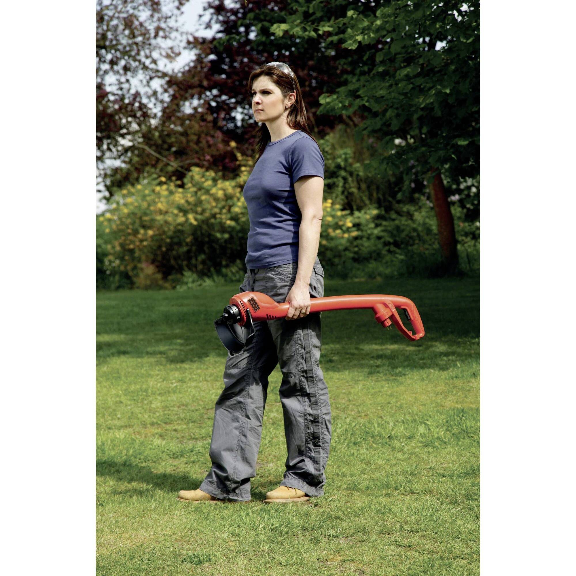 A woman is standing on a lawn and holding a red lawn trimmer in her right hand. Trees can be seen in the background.