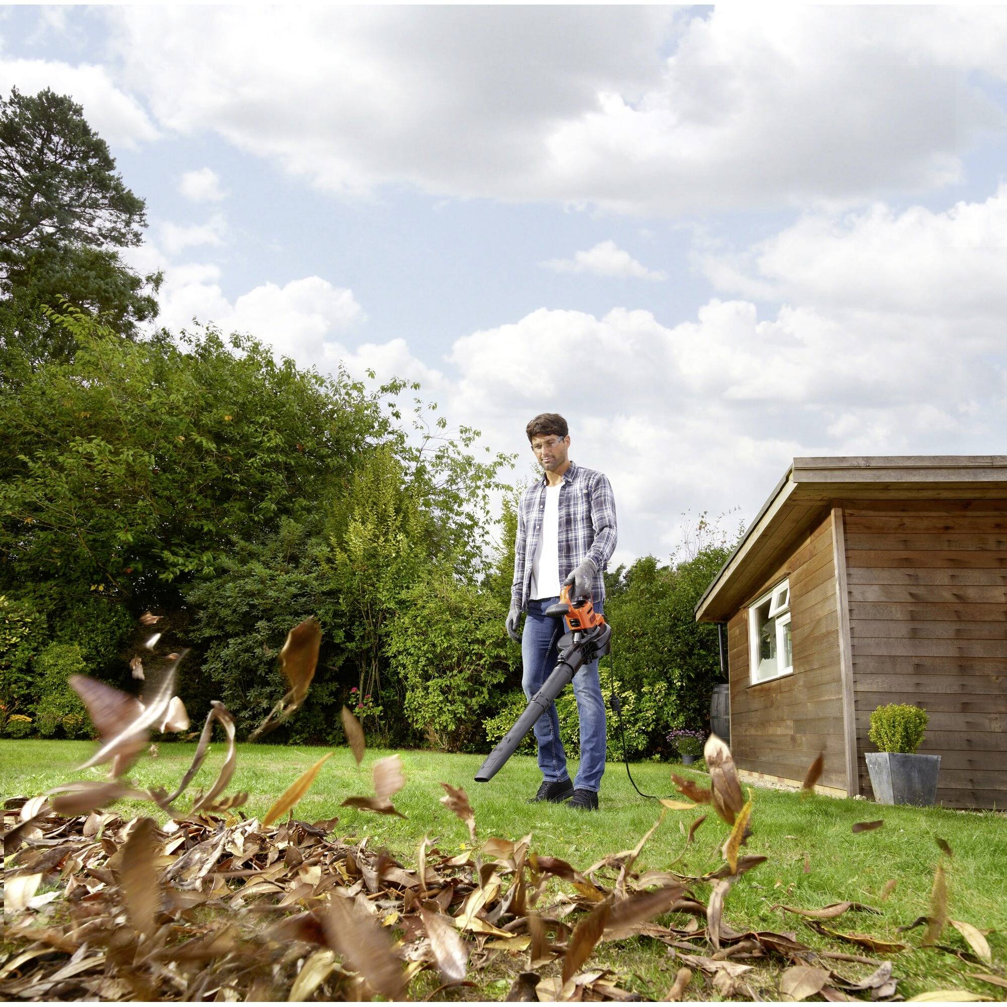 A man is using a leaf blower in the garden next to a wooden house. Leaves are piling up in the foreground, with a tree visible in the background.