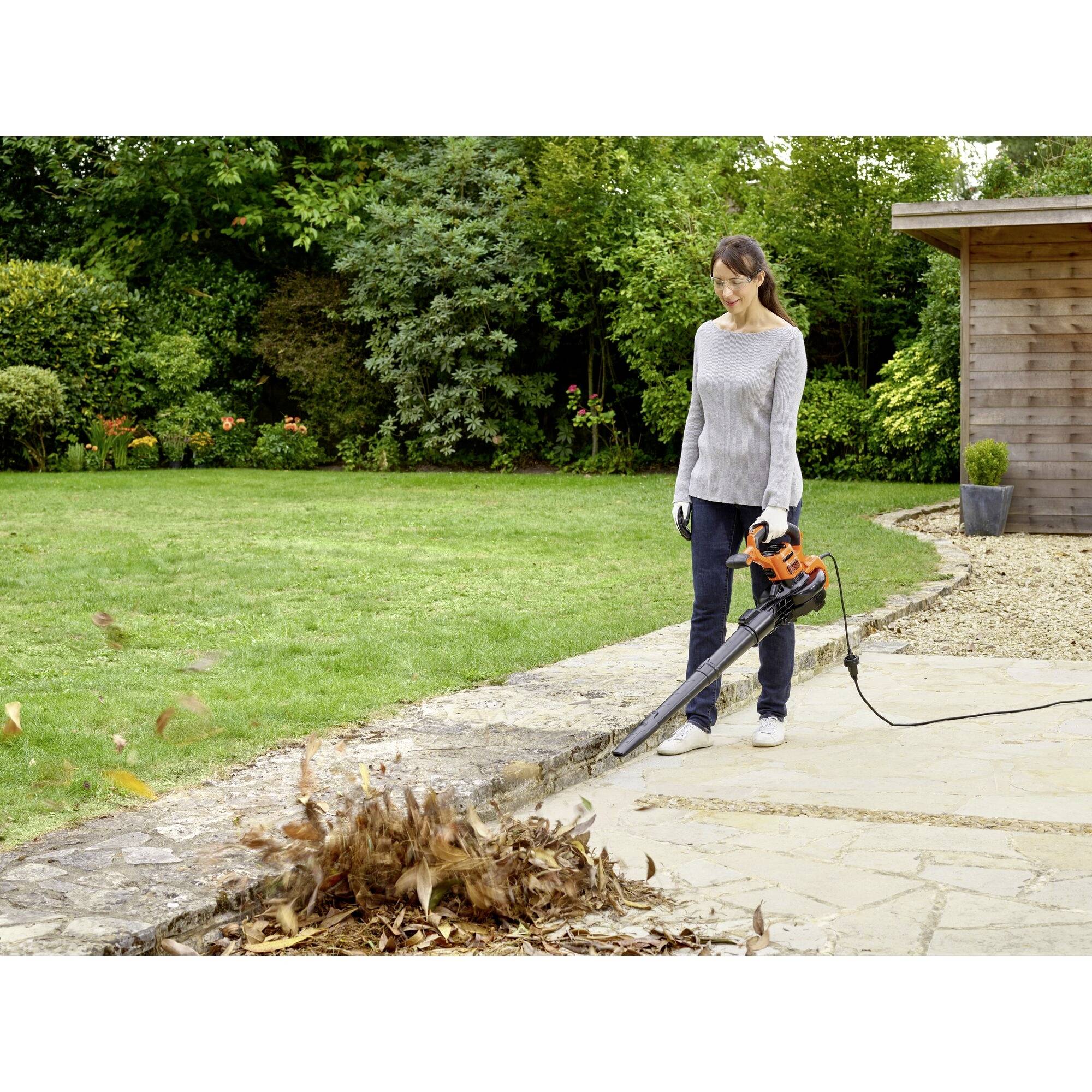 A person is using a leaf blower to blow leaves from a patio into a garden. Trees and a shed are visible in the background.