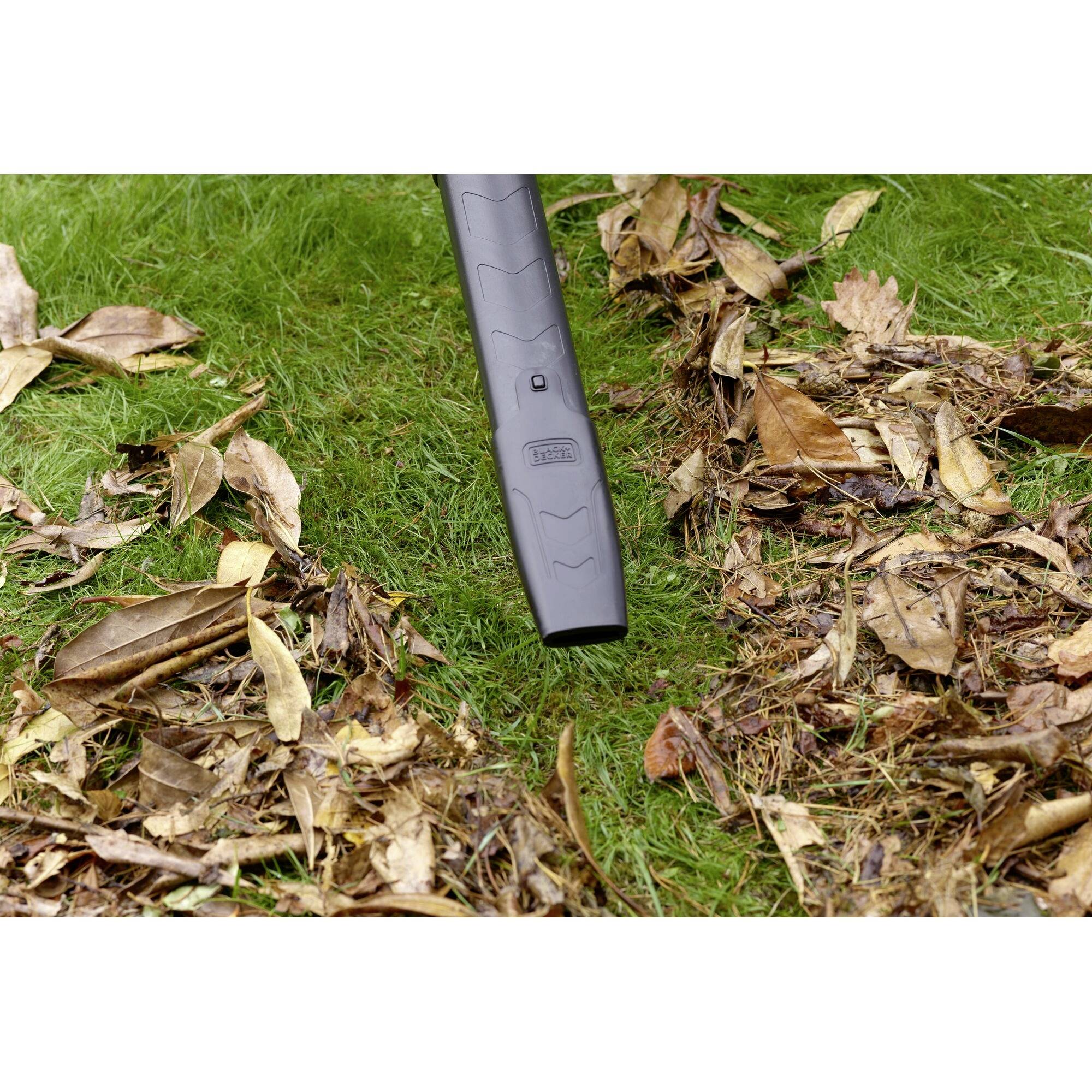 A leaf blower is sucking dry leaves from a lawn. The ground is covered with green grass and brown leaves.