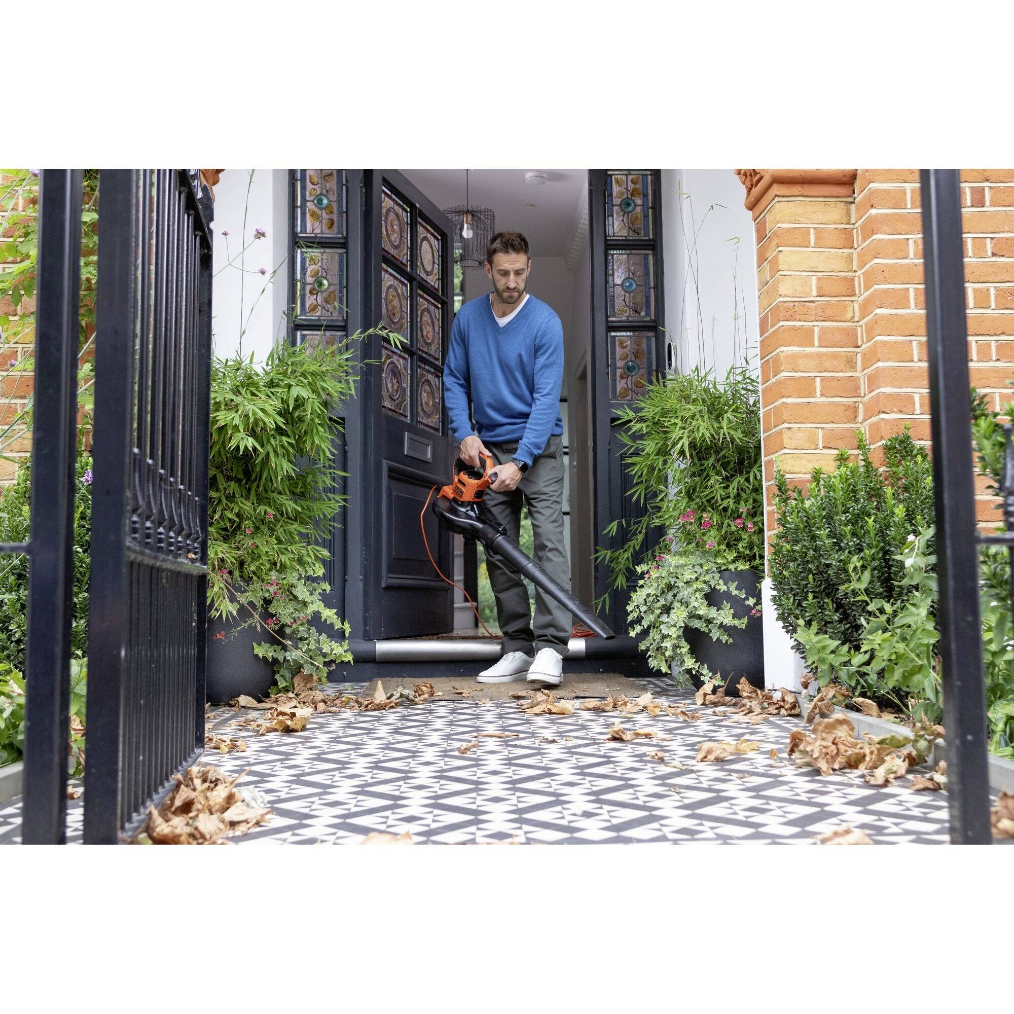 A man is standing on a tiled terrace with a leaf blower in his hand, surrounded by plants and colourful foliage.