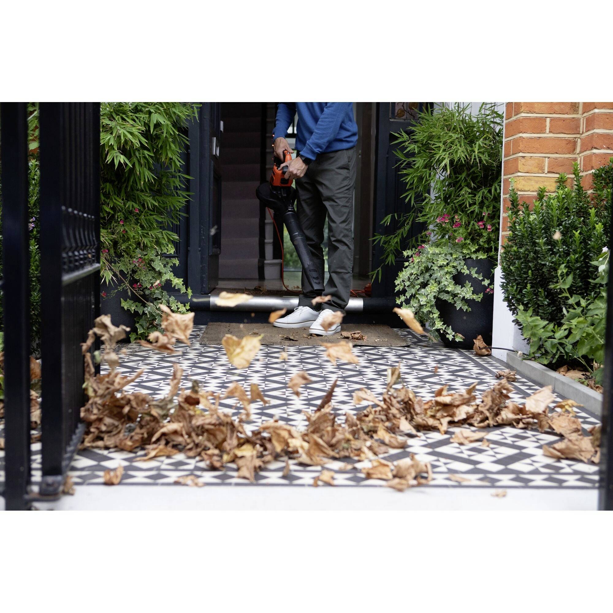 A person is blowing dry leaves with a leaf blower on a patterned floor near an entrance, surrounded by green plants.