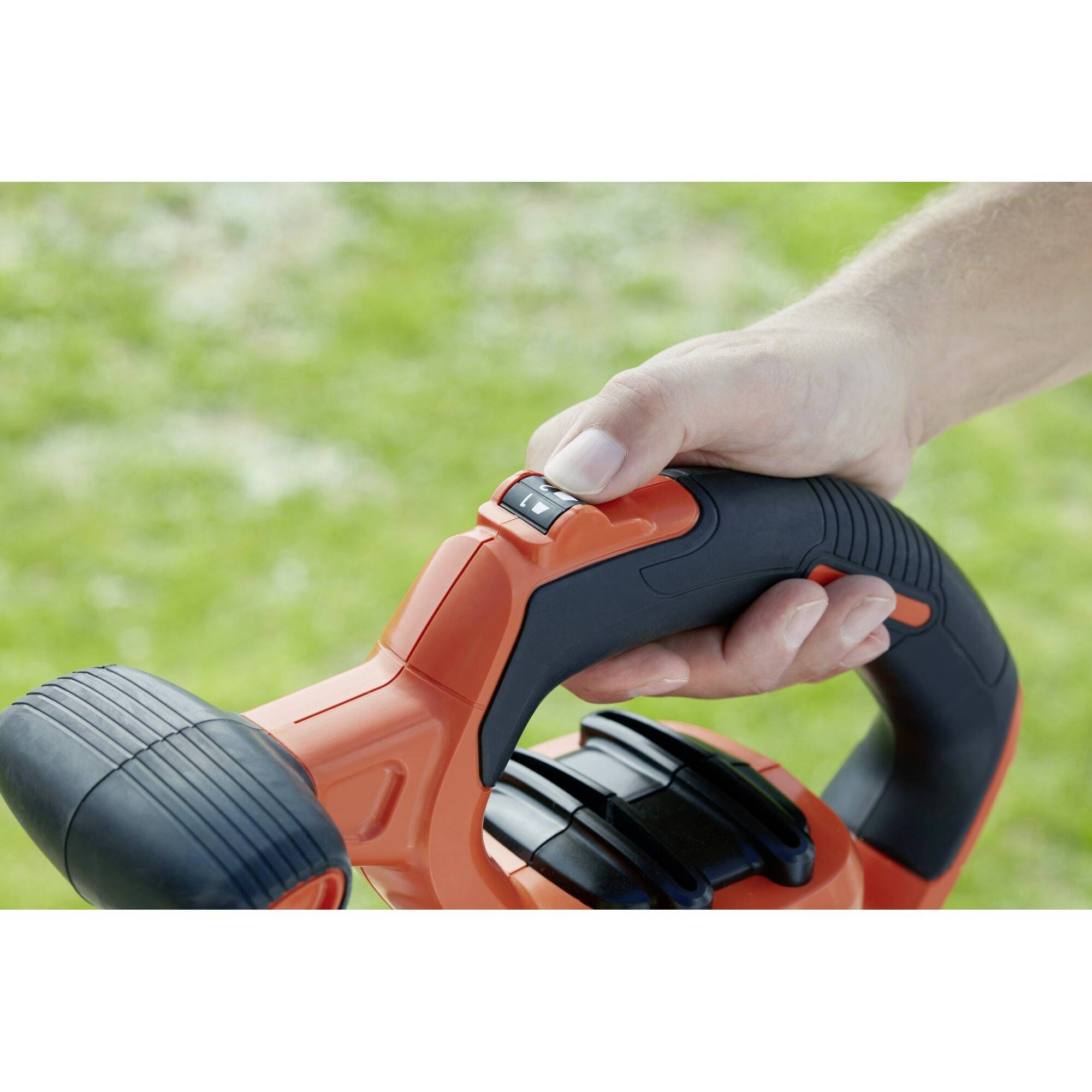 A hand is pressing the black handle of an orange lawn mower trimmer with a small control button outdoors.