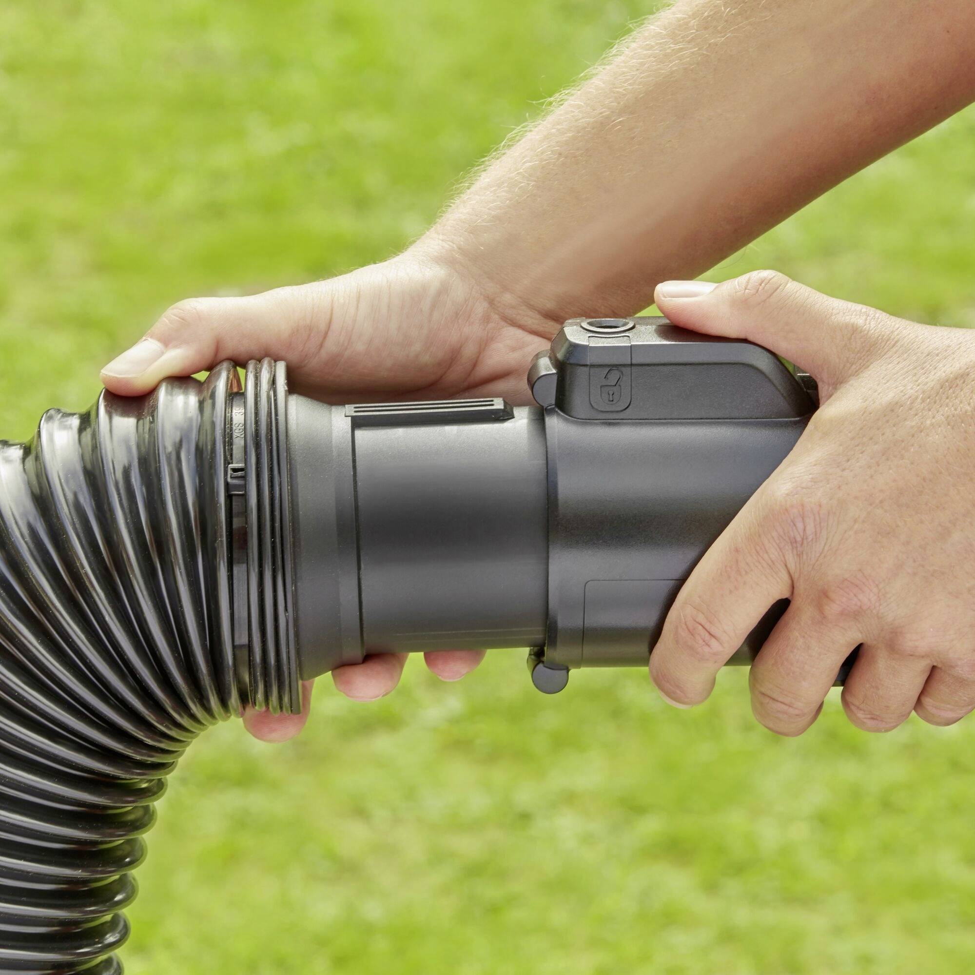 A man connects a black hose with another plastic component on green grass. The focus is on the hands and the hose.