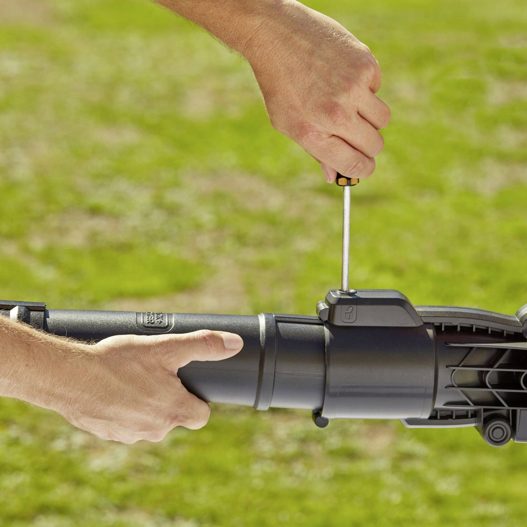 A person is securing a part to a garden sprayer with a screwdriver. Hands are in focus, with a green background suggesting an outdoor setting.