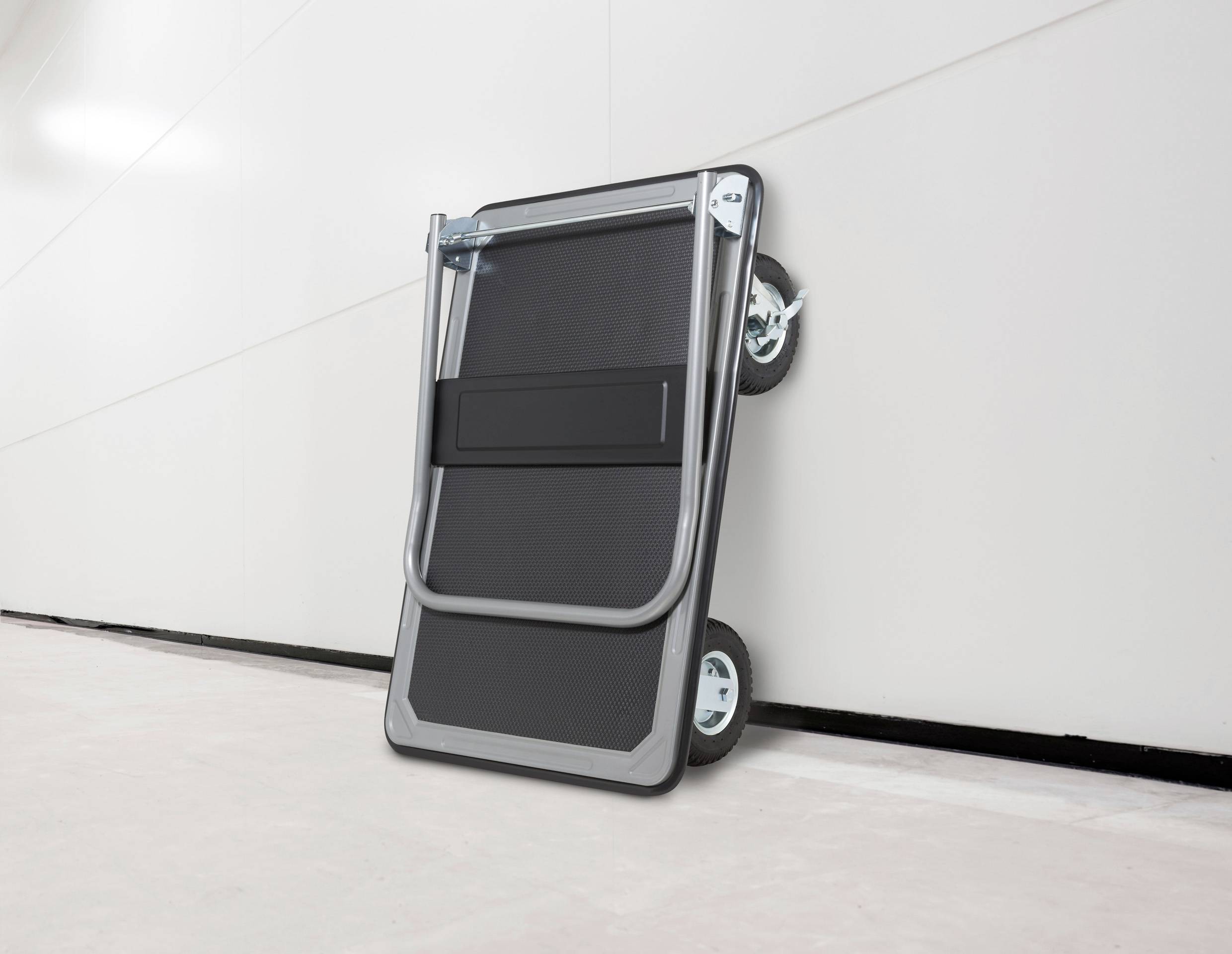 A folded hand truck with two wheels leans against a smooth, white wall on a concrete floor.
