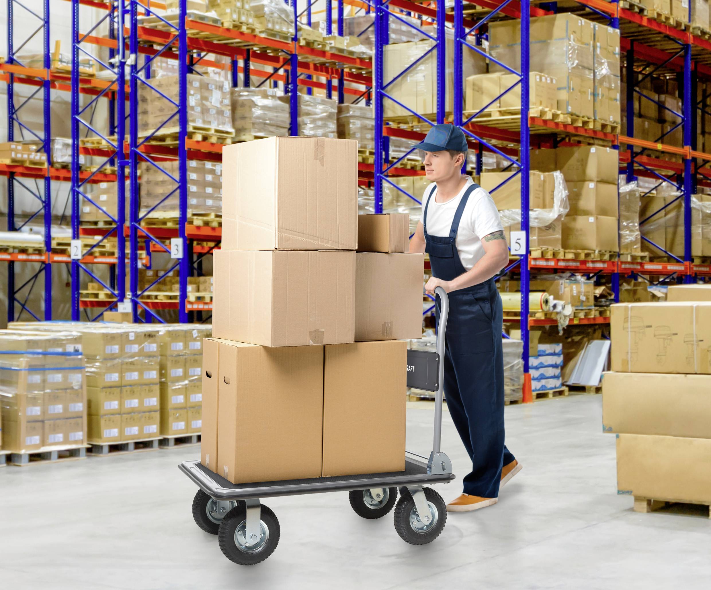 A person wearing overalls is pushing a cart loaded with large cardboard boxes in a warehouse filled with shelves of various packages.