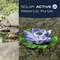 Solar-powered water lily in purple floats on water. Background shows stones and grass. Text: 'Solar Active WaterLily Purple'.