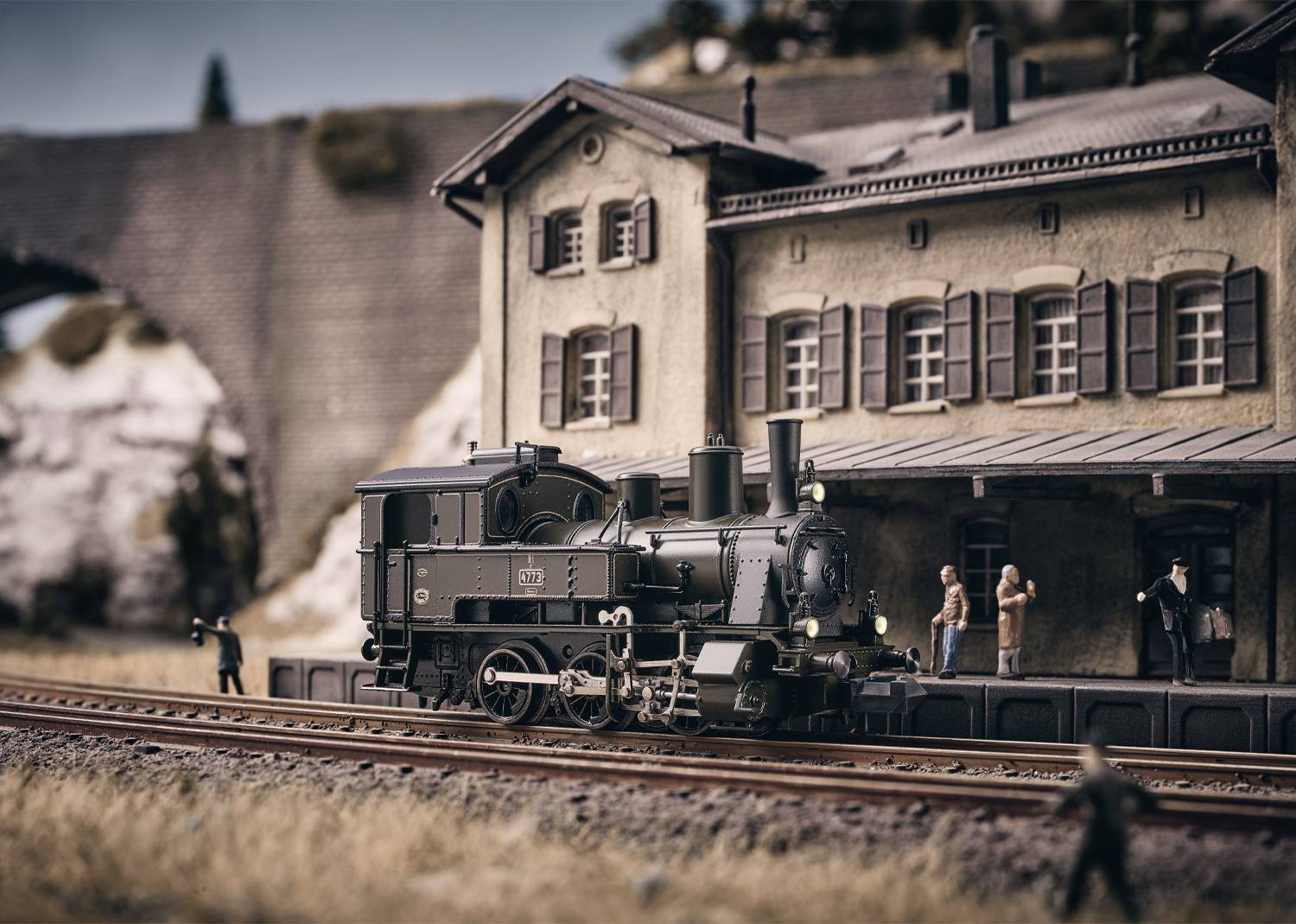 Model railway with a historic steam train in front of an old railway station. Several figures are standing on the platform. The background shows a stone bridge.