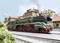 Green and red steam train travels on track past historic buildings under blue sky. Smoke rises from the chimney.