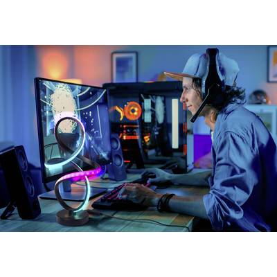 A person wearing a headset plays a video game on a computer, surrounded by a colorful, illuminated gaming setup in a dimly lit room.