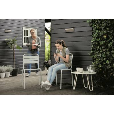 A woman sits outside on a chair and looks at her phone. Another person comes out of a building with a tray.