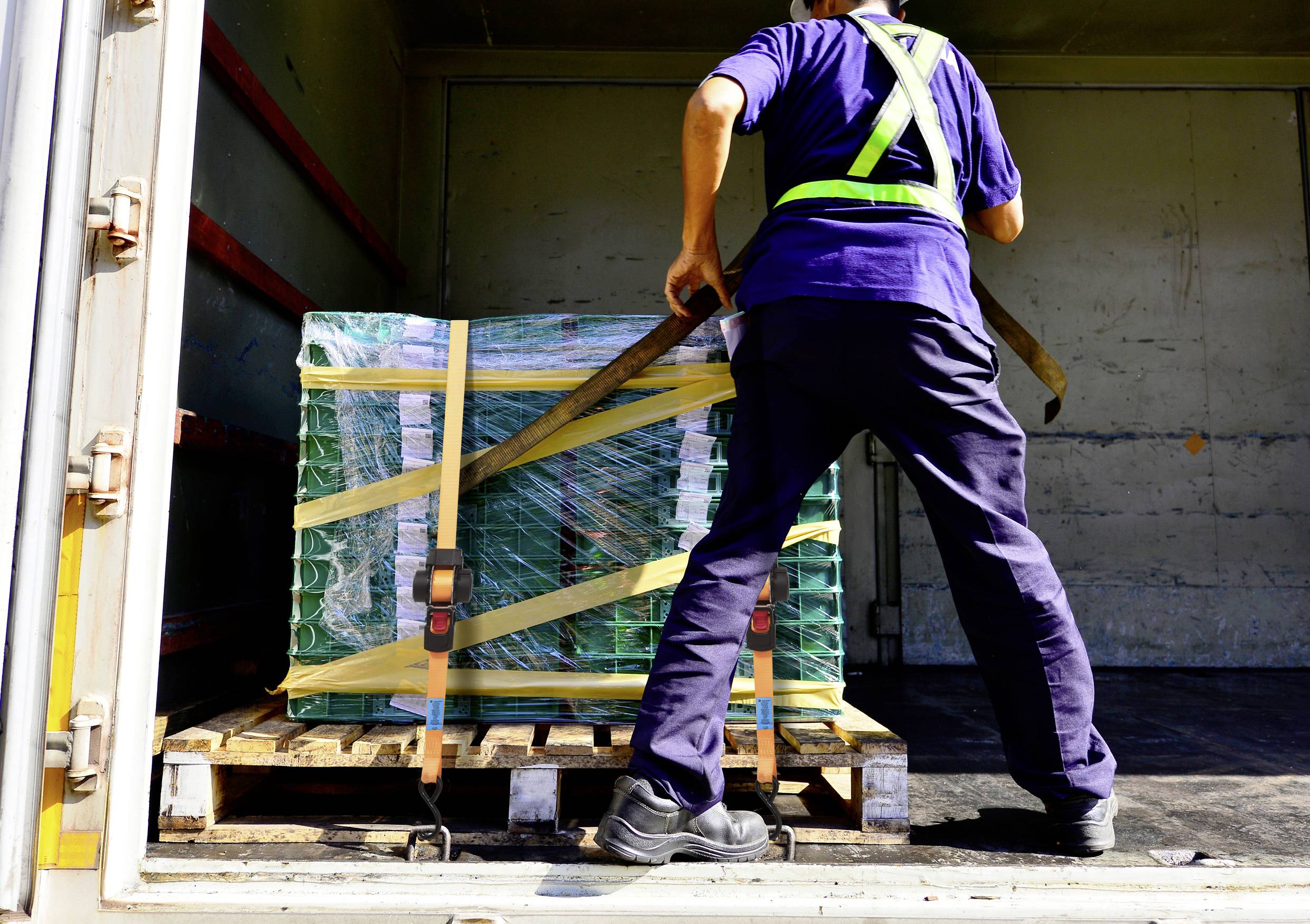 A worker loads a large, green-wrapped pallet onto a truck using a pallet jack, wearing a hi-vis safety harness and uniform.