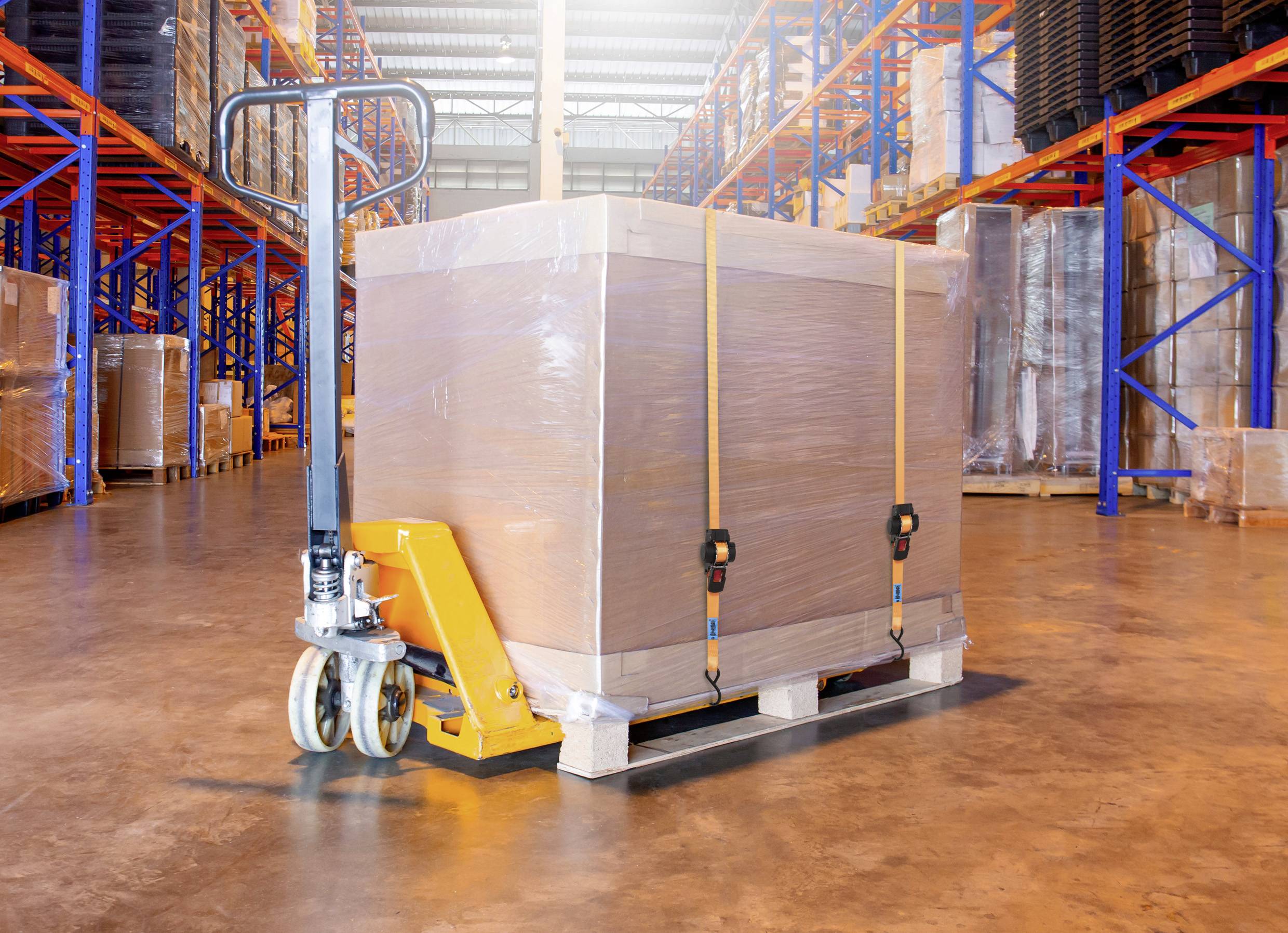 A warehouse with a pallet jack transporting a large, wrapped package on a wooden pallet. Shelves with stacked goods are visible in the background.