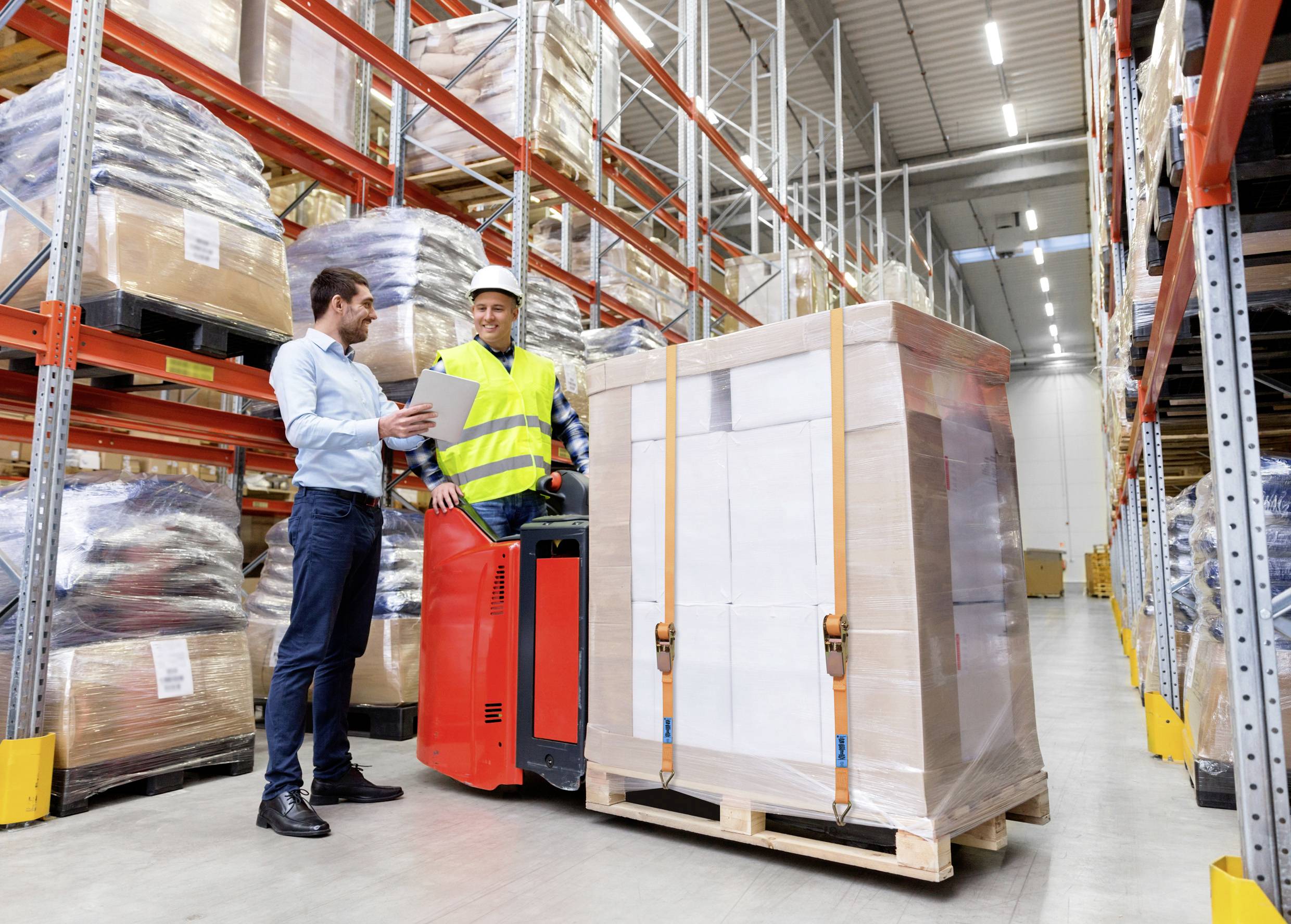 A warehouse worker operates a pallet jack loaded with boxes, while a person holds a clipboard, suggesting a shipment review.