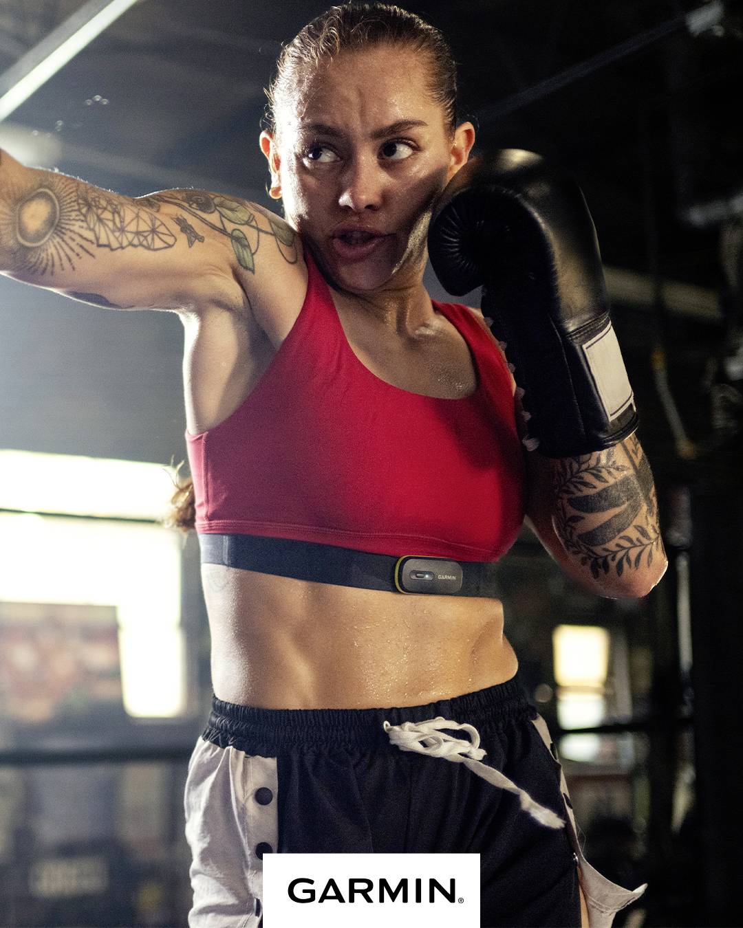 A woman during boxing training wearing a red sports bra, dark gloves, and with tattooed arms. The Garmin logo is visible in the lower area.