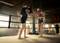 Two people are training boxing in the ring. One person is wearing boxing gloves, the other is holding focus mitts. Boxing bags are visible in the background.