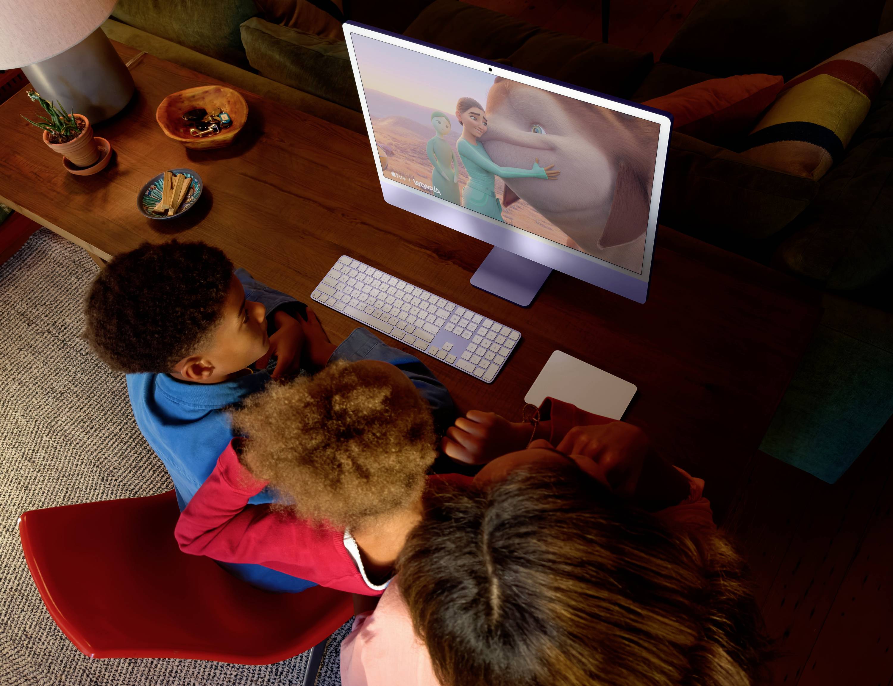 A family of three watches an animated movie on a computer screen in a cozy living room, surrounded by cushions and plants.