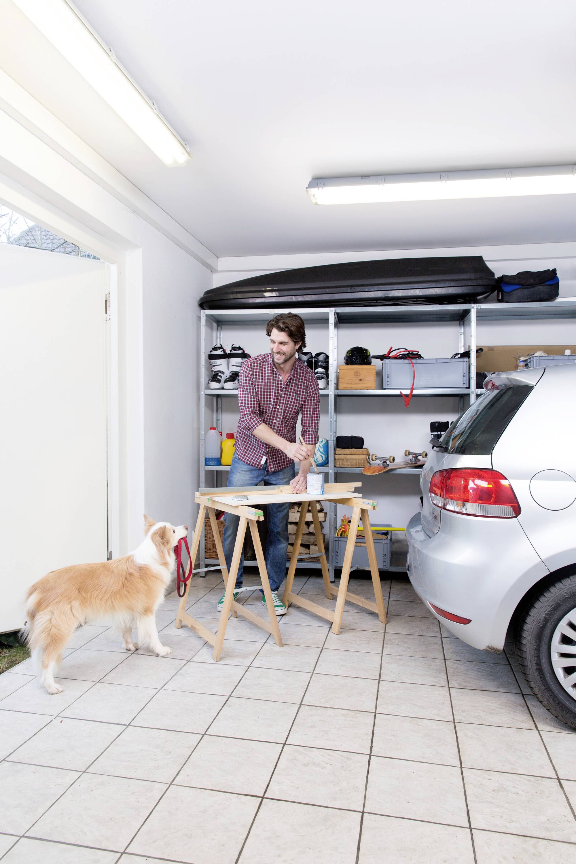 A man is working at a workbench in a garage while a dog watches. A car is parked nearby.