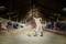 A person in work clothes is sweeping the floor of a cattle barn, with cows visible on the left and right sides.