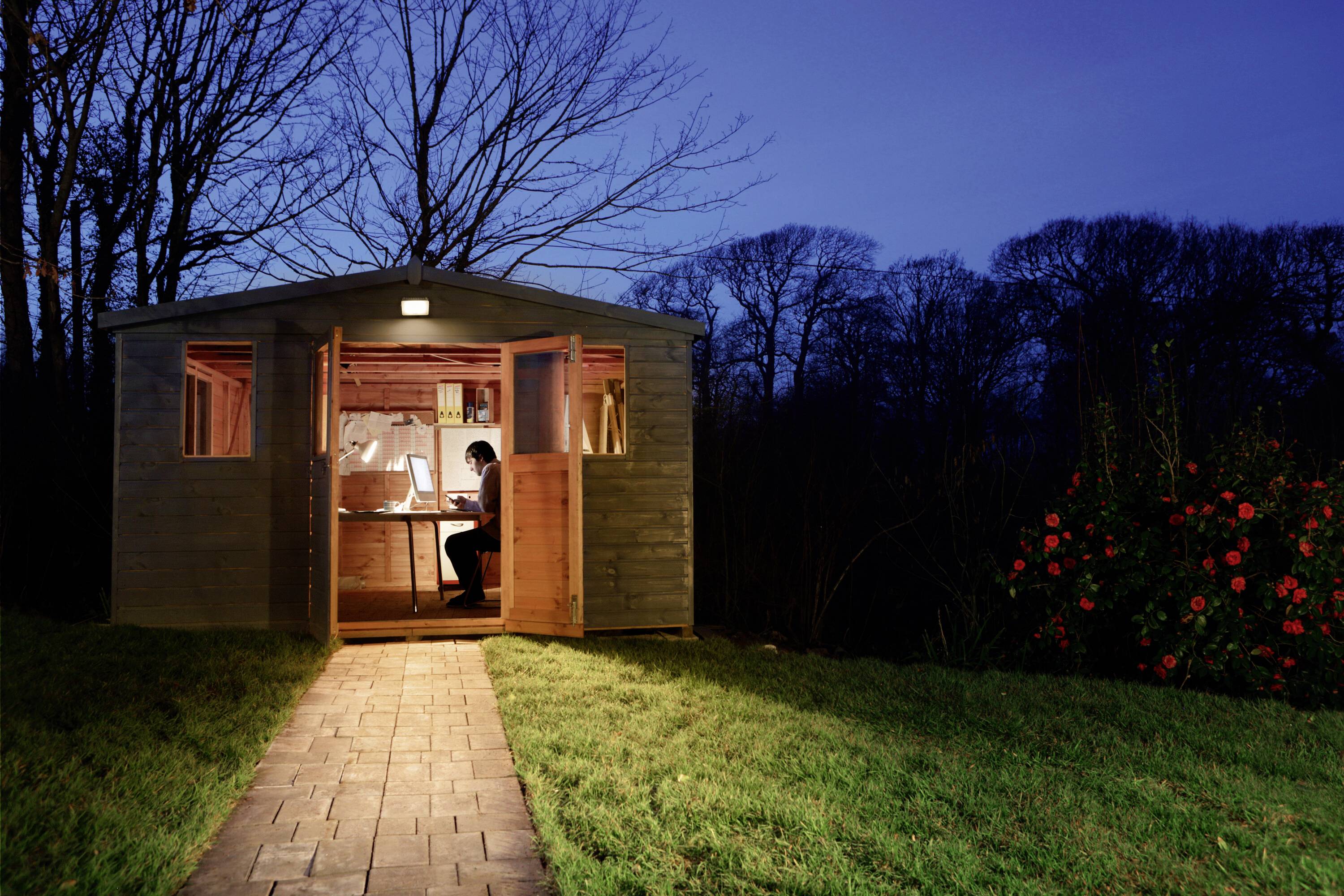 A person is working in the evening in an illuminated garden house, surrounded by trees and a blooming bush.