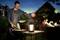 A man is grilling in the garden in the evening, while a group sits at a table in the background. Lanterns are hanging, and it looks like a sociable celebration.