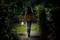 A person walks at night on a narrow garden path, holding an illuminated lantern that gently lights the way.