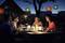 Four people are sitting at an outdoor table, smiling and chatting by candlelight. Colorful lanterns hang above them.