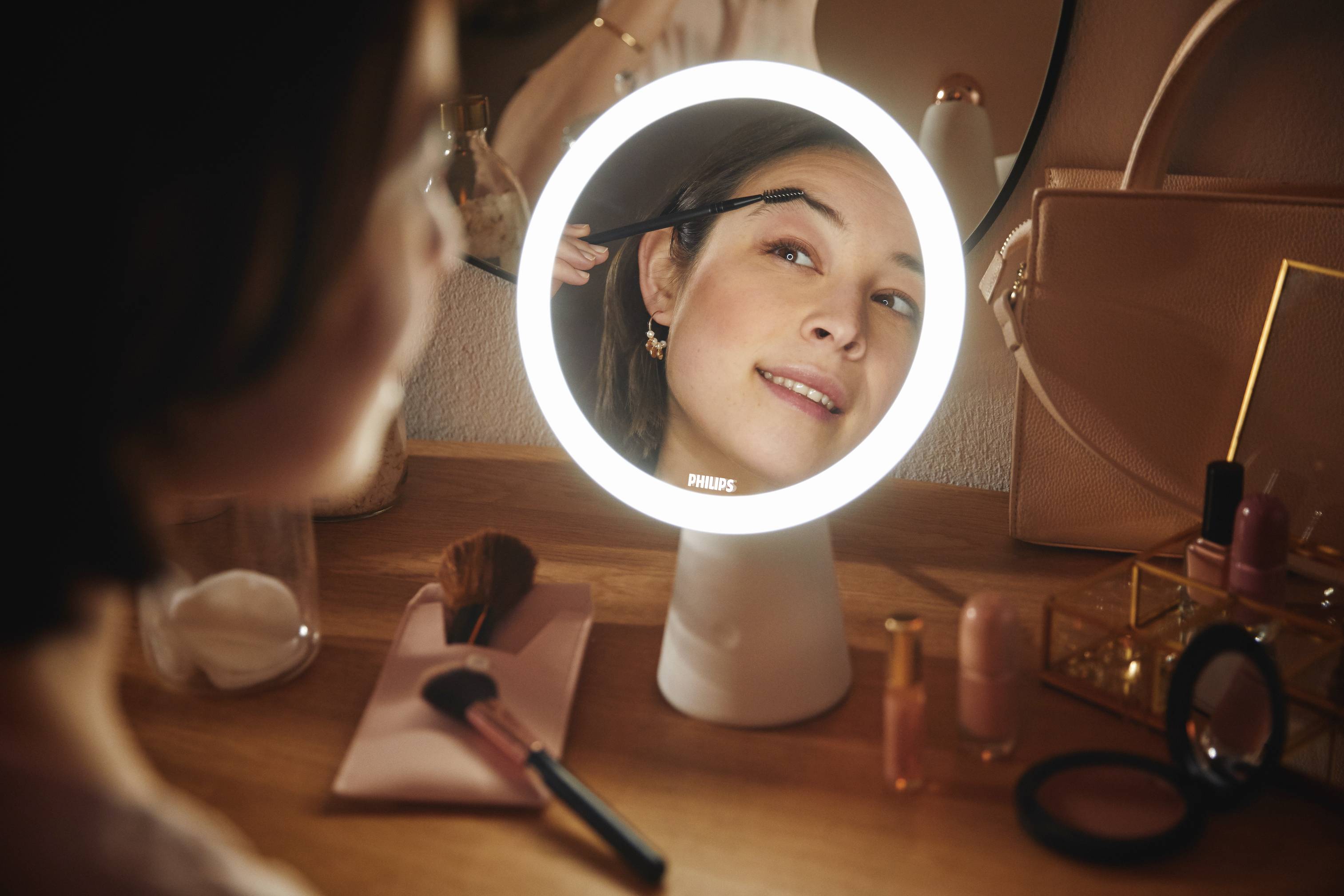 A woman is applying makeup in front of an illuminated mirror. Makeup utensils are lying on the table.