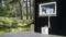 A portable air conditioning unit stands on a wooden deck in front of a black house with a wooded background. A hose leads to the window.