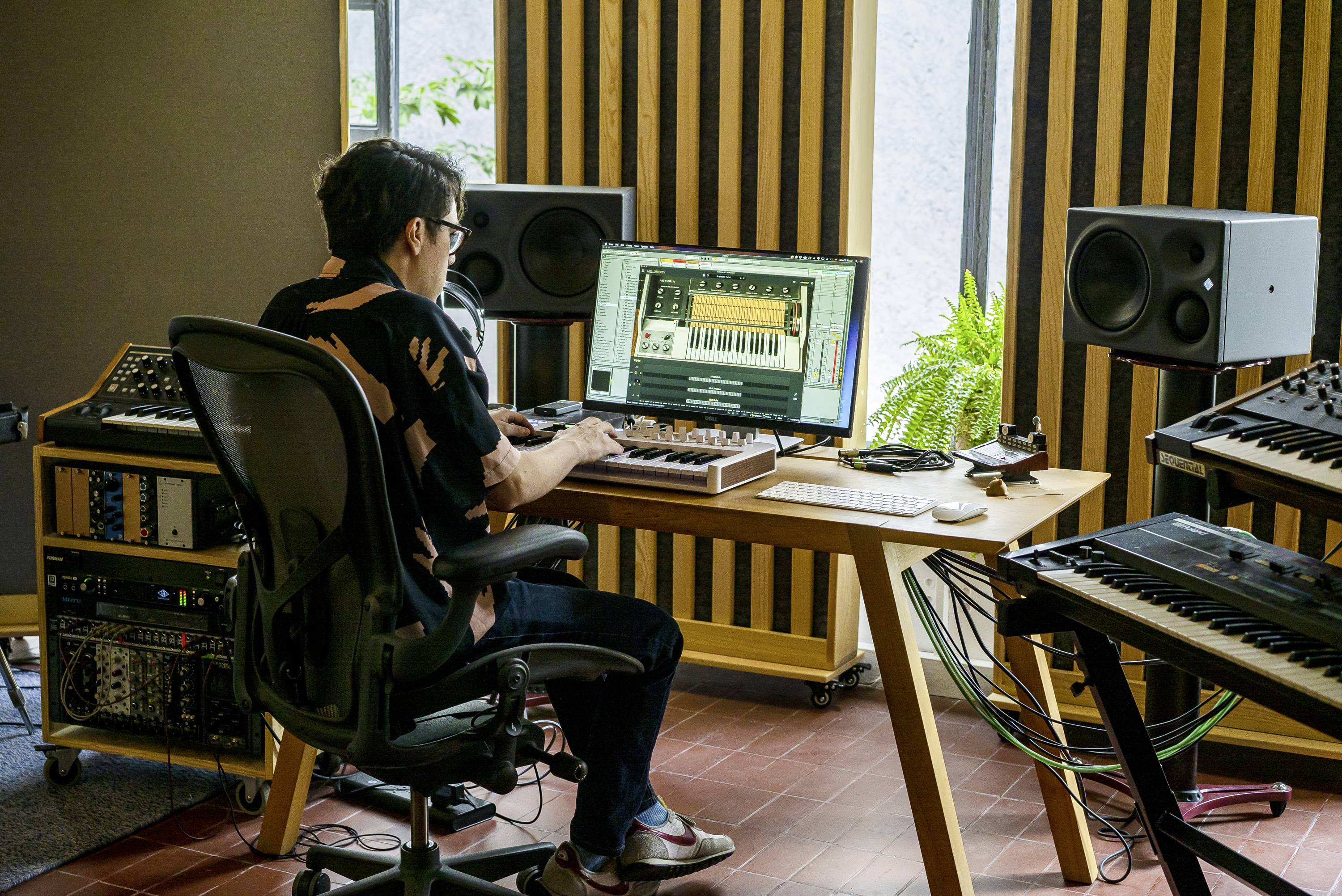 A person sits at a desk in a music studio, working on a music program on a computer, surrounded by synthesizers and speakers.