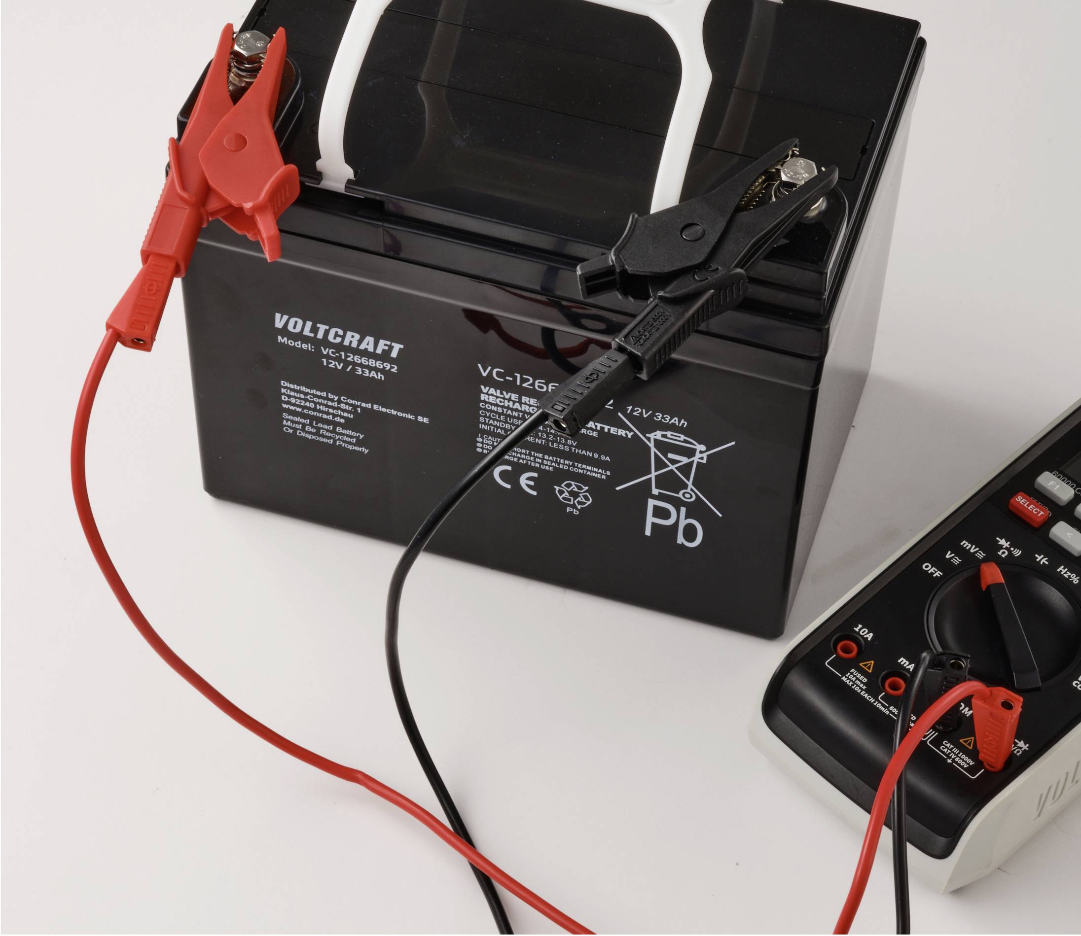 A car battery with red and black jumper cables connected, and a digital multimeter measuring voltage.