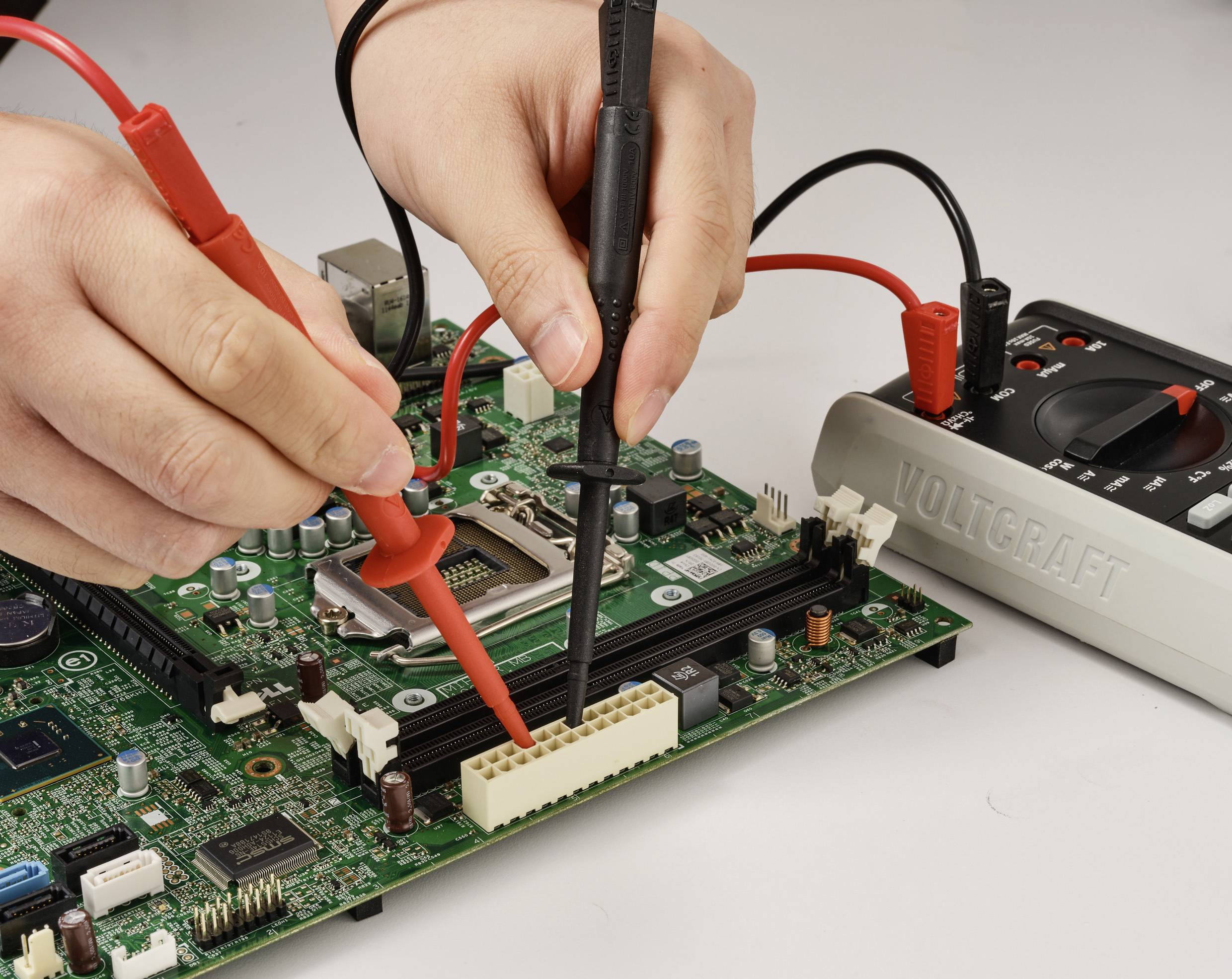 Hands using multimeter probes to test a circuit board. The red and black probes are connected to different points on the board.