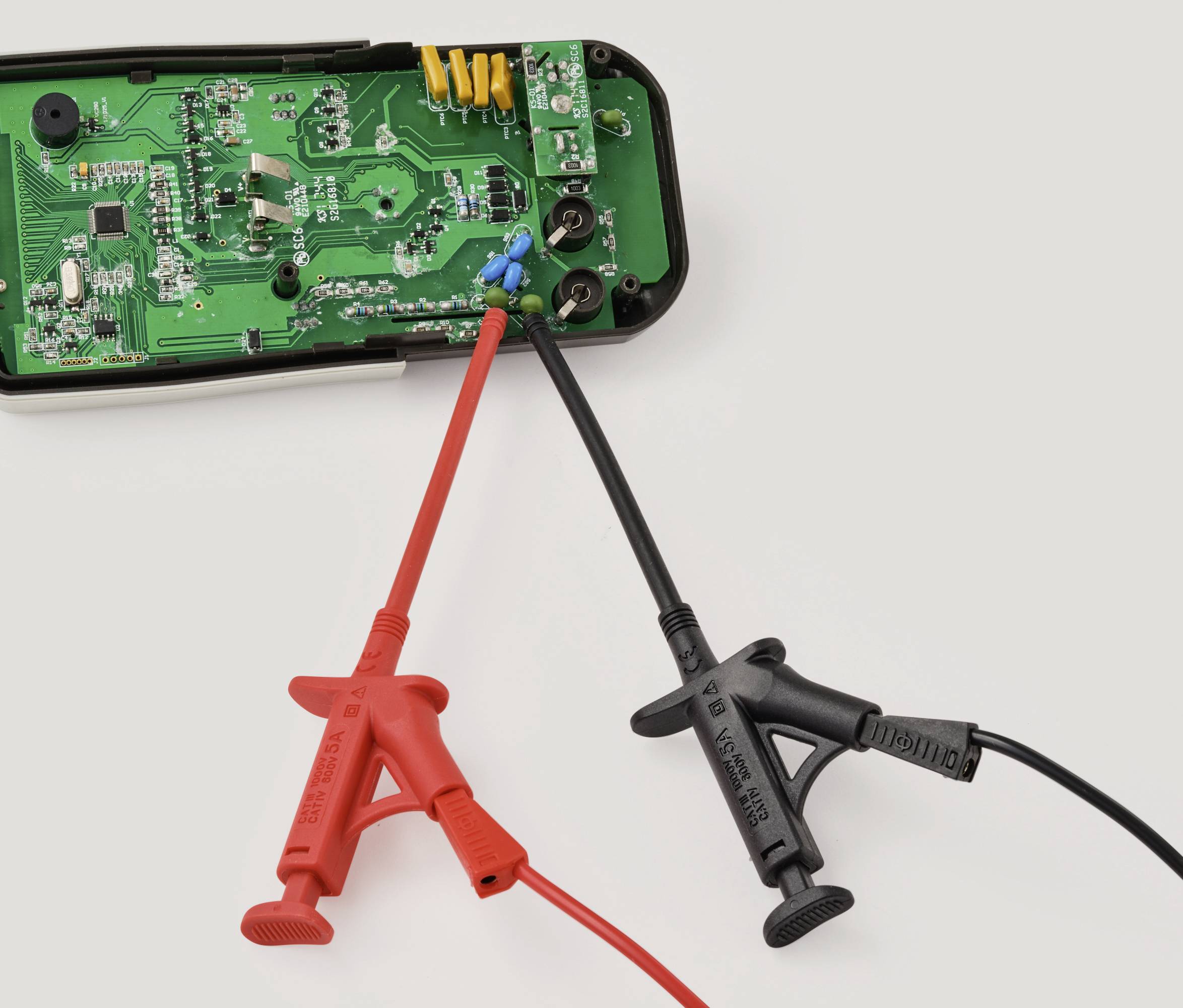 A green circuit board with electronic components is connected to a red and black test probe. The device's purpose involves electrical testing.