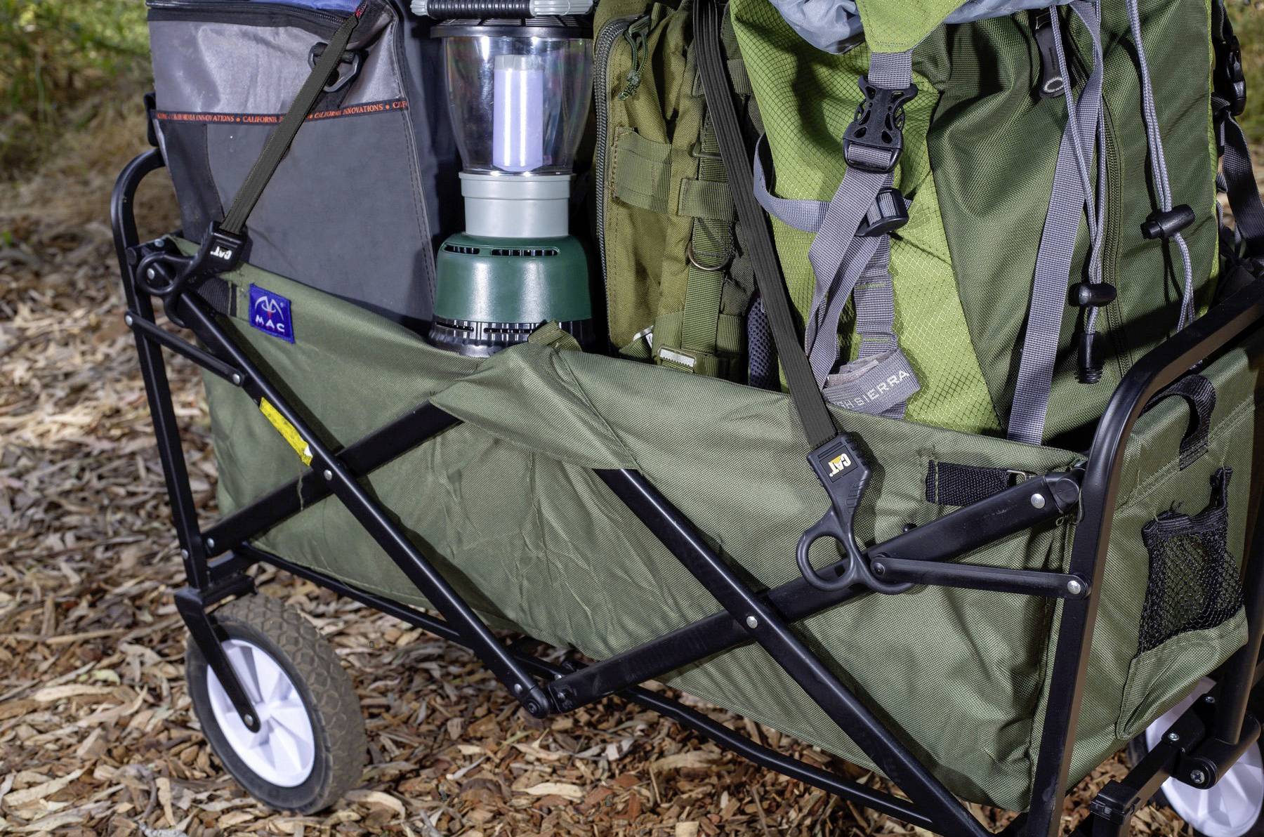 A cart filled with camping gear including backpacks, a lantern, and pouches on a leafy ground outdoors.