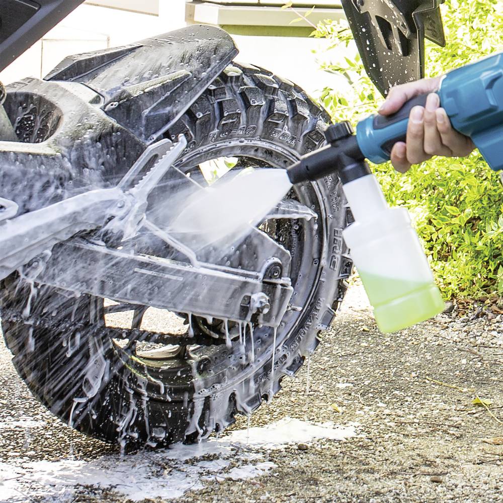 A person uses a pressure washer with soap to clean a motorcycle tire. The foam covers the wheel, indicating thorough cleaning.