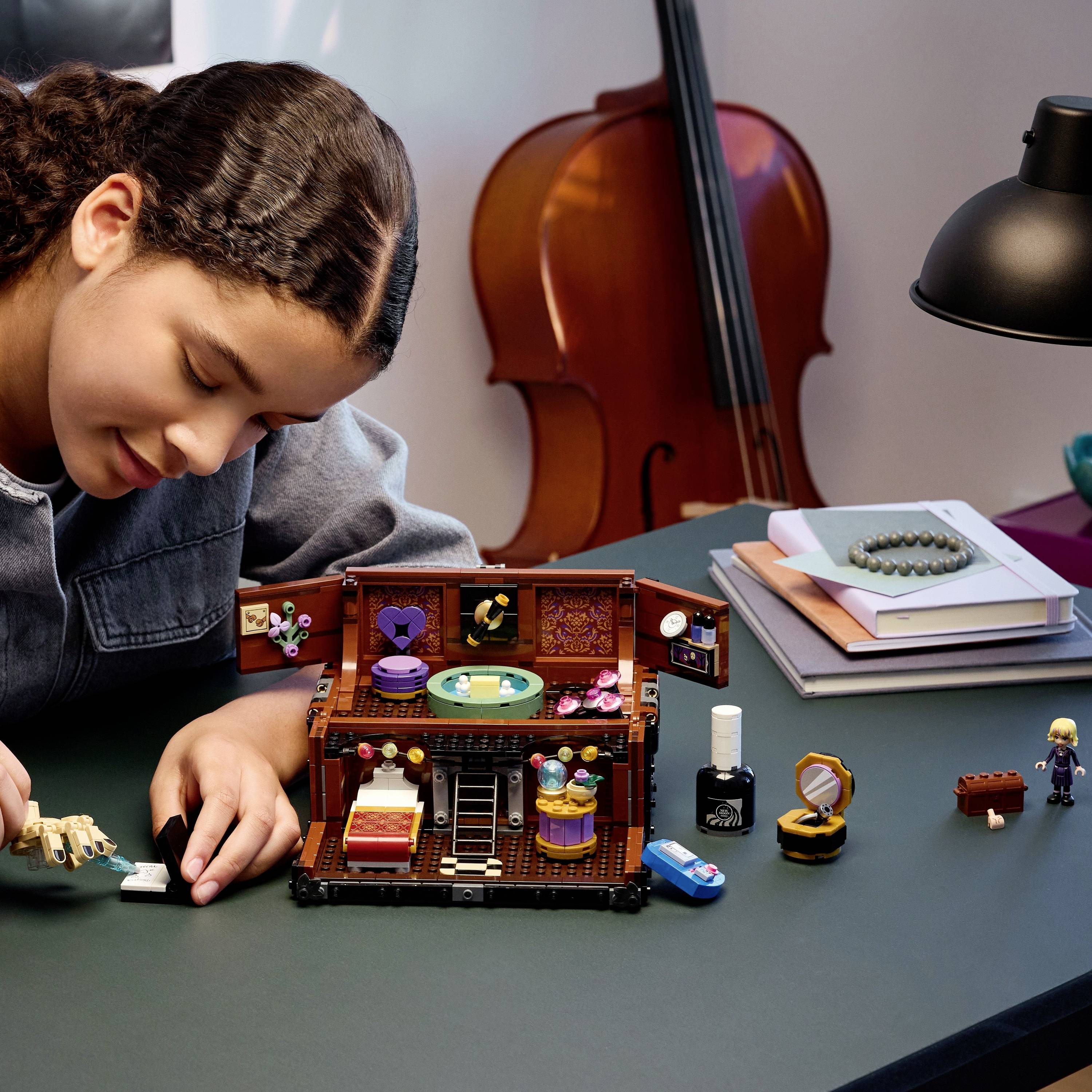 A person builds a small, detailed model scene with furniture and accessories on a table, surrounded by craft supplies, a lamp, and books.
