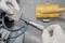 A person wearing gloves uses a micrometer to measure a metal block on a workbench, with a yellow tool case visible in the background.