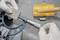 A person measures a metal block labeled 'S5 1.2' with a micrometer caliper. Tools are on the table, including a yellow case.