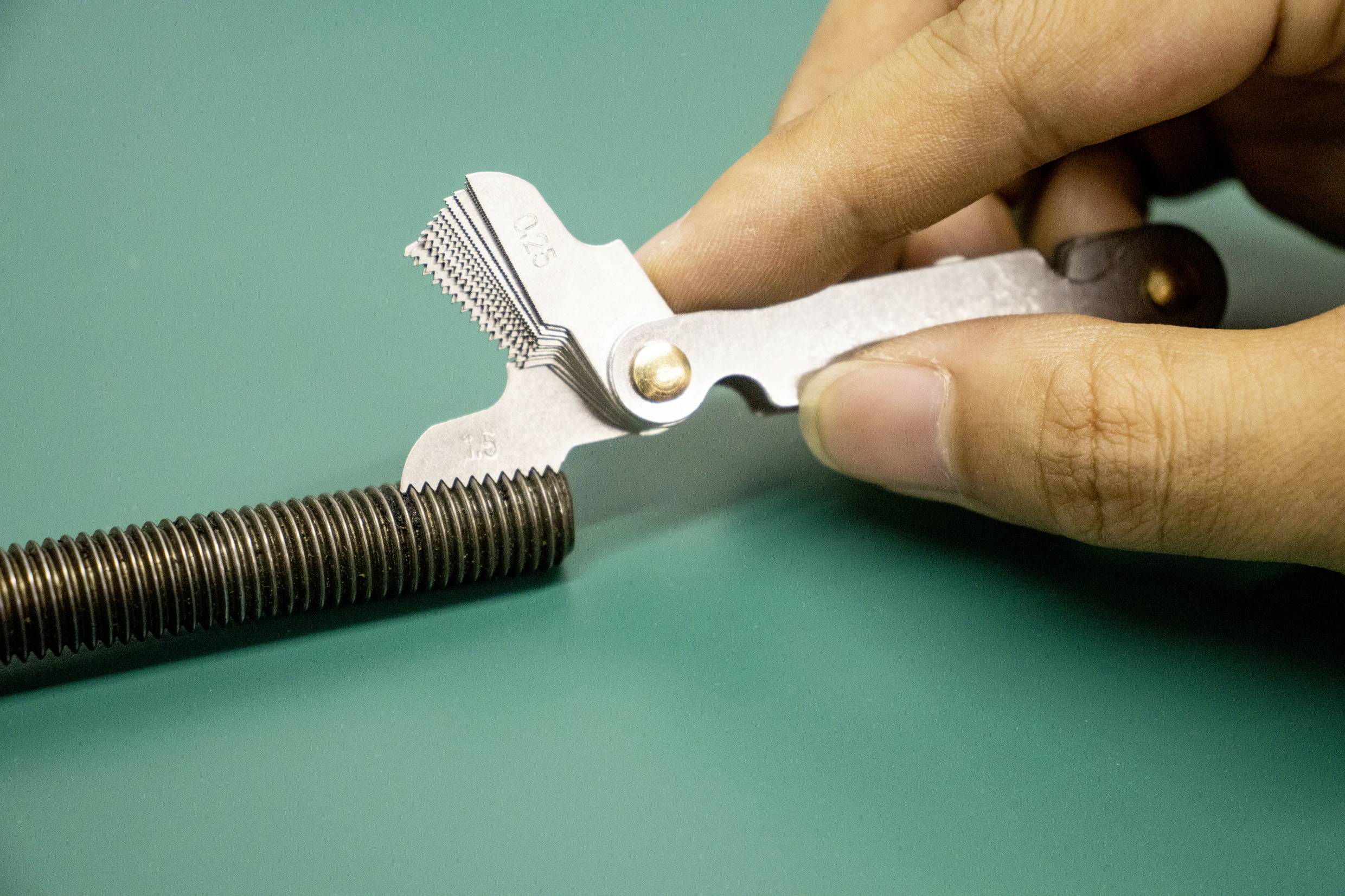 A person uses a thread gauge to measure the pitch of a metal screw against a teal background.